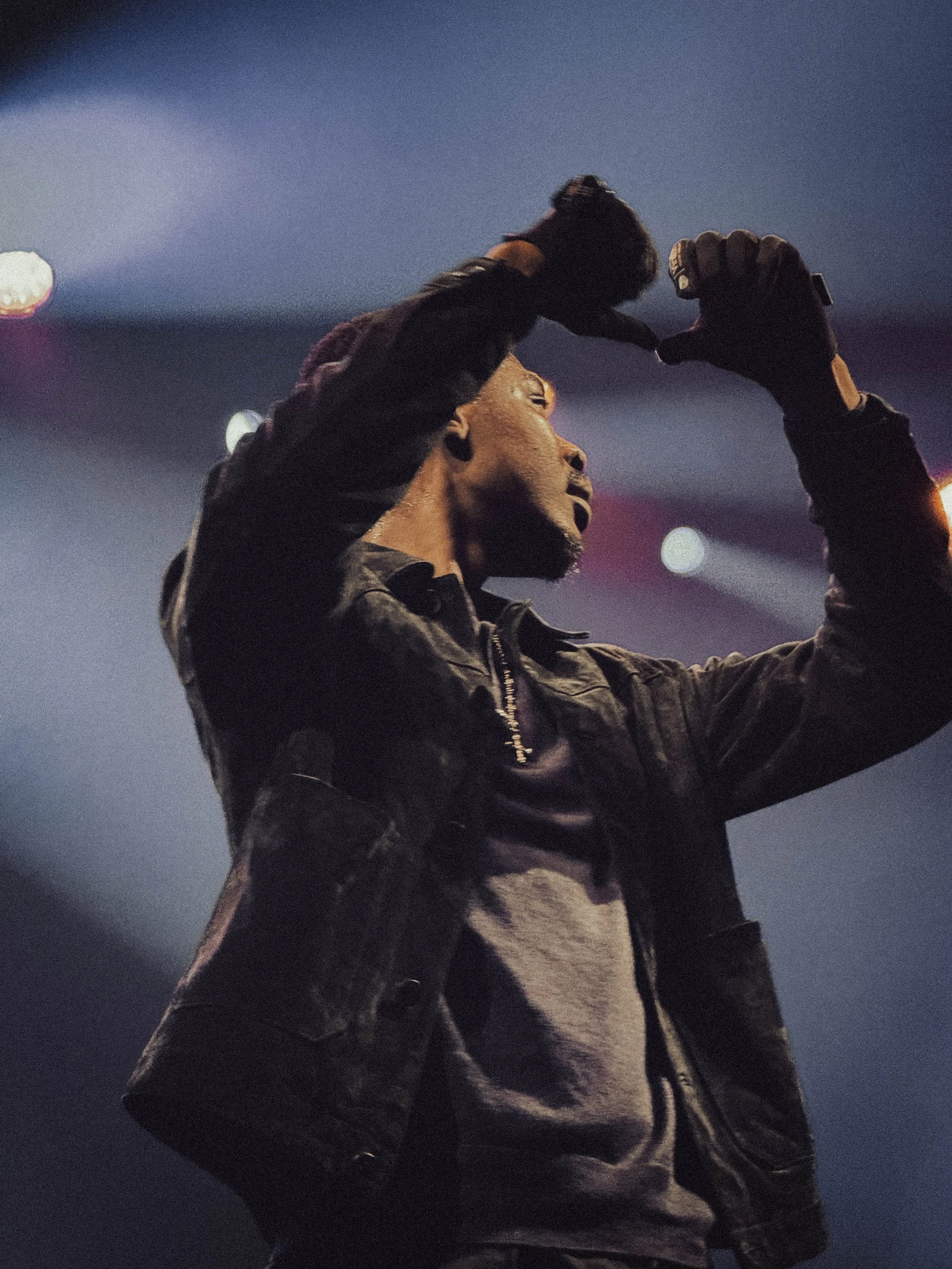 A man wearing a black jacket and gloves, with a beard, is dancing or performing on stage against a dark background with blurred lights.