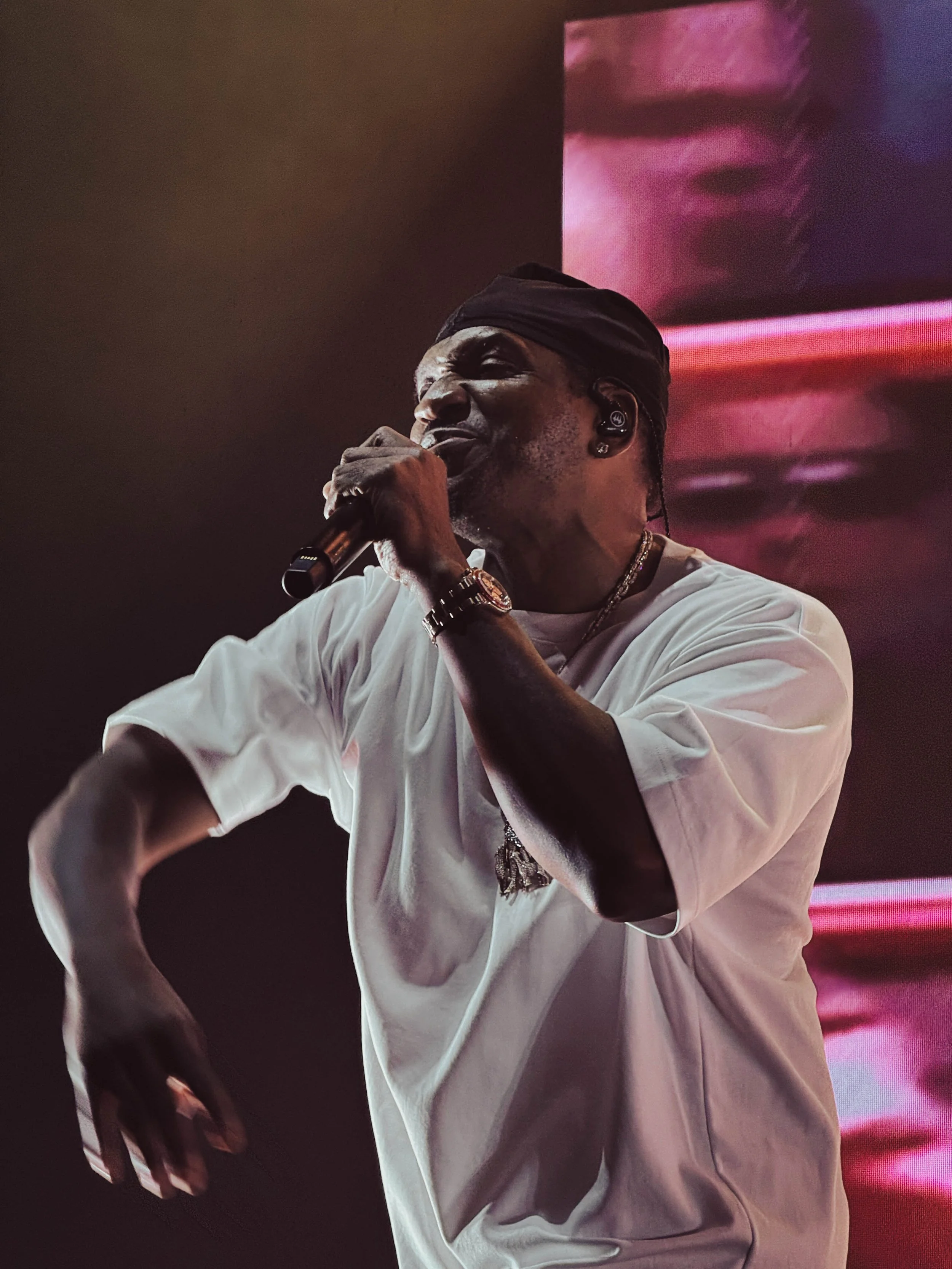 A male singer wearing a black headband, white oversized T-shirt, and jewelry, passionately performing with a microphone on stage with a large digital screen displaying a distorted face in the background.