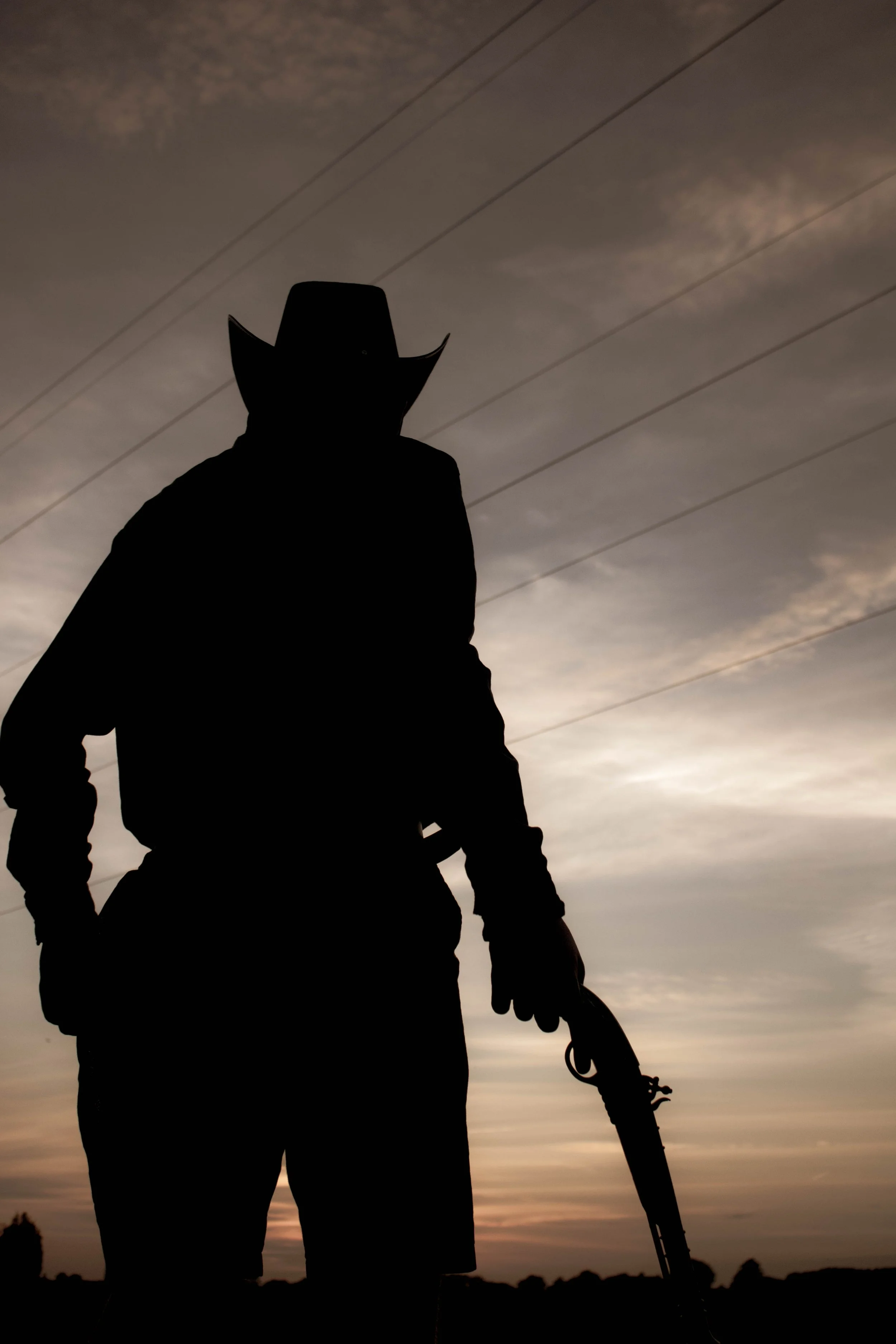 Silhouette of a person wearing a cowboy hat holding a rifle, standing outdoors during sunset or sunrise with a cloudy sky and power lines in the background.