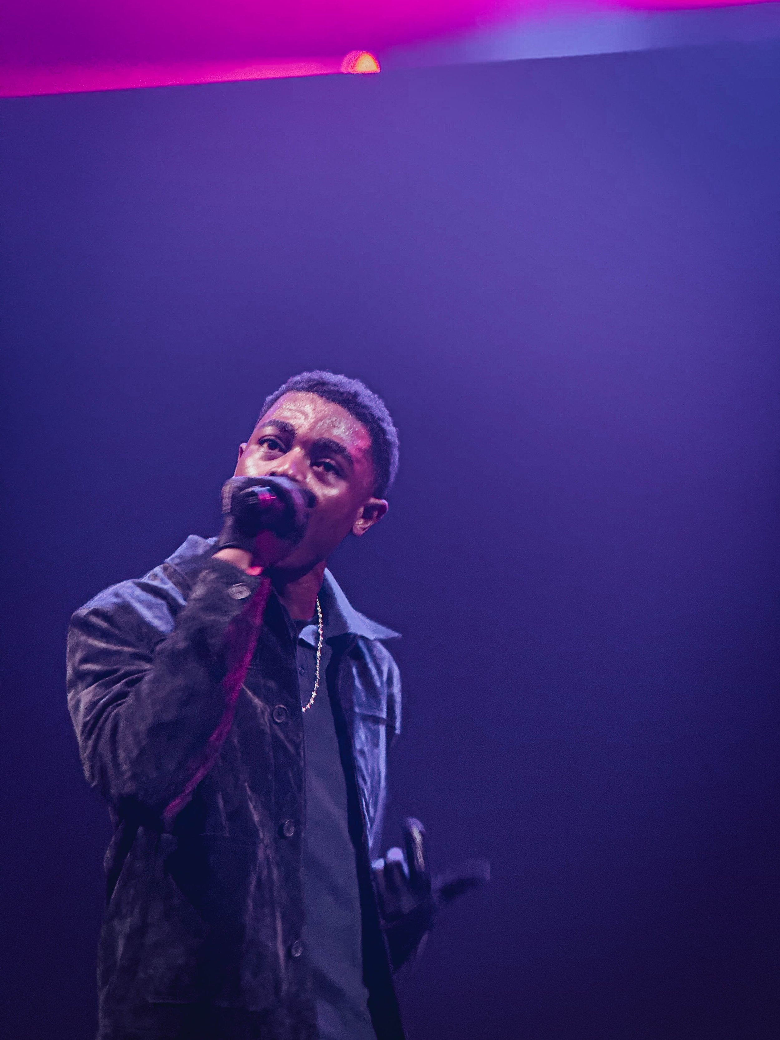 Young man holding microphone on stage under purple and pink lighting.