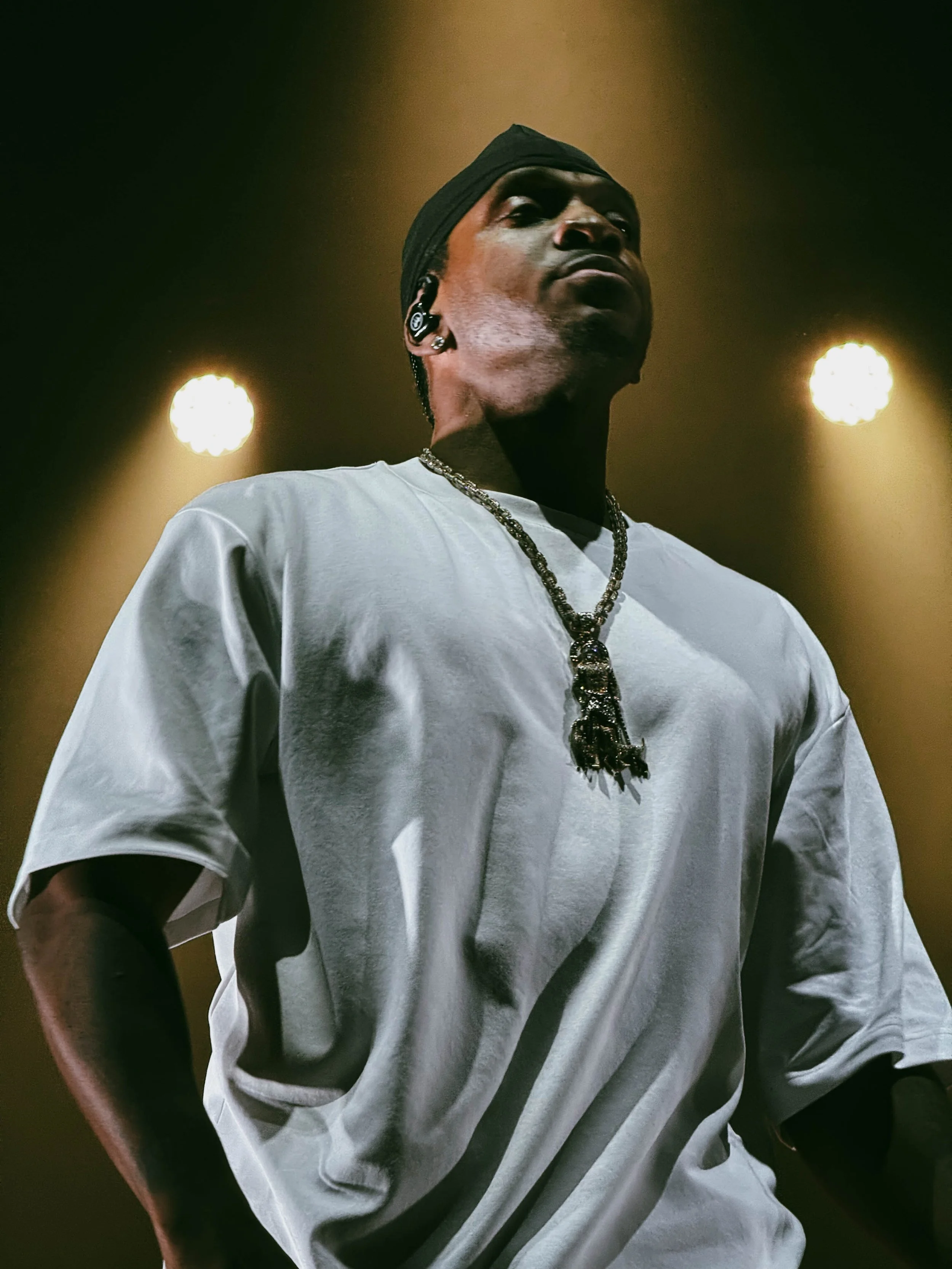A man wearing a black head covering, a white t-shirt, a beaded necklace, and earrings, standing on stage with yellow backlighting.