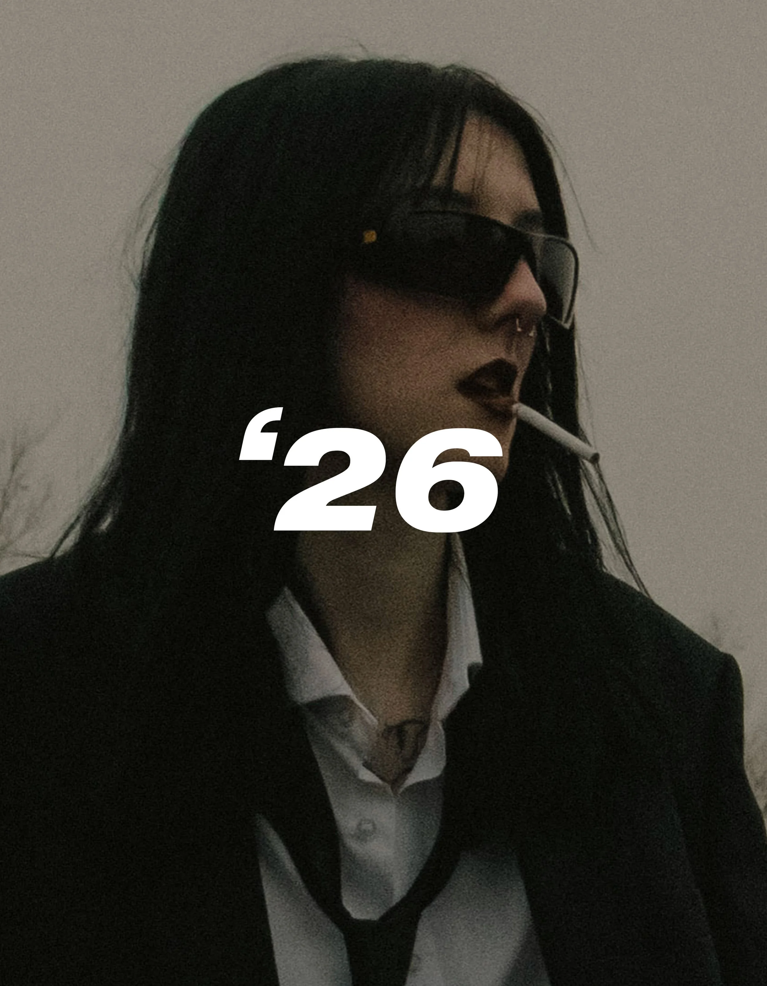 A woman wearing black sunglasses with a cigarette in her mouth, dressed in a black blazer over a white shirt, with '26' written across her face.