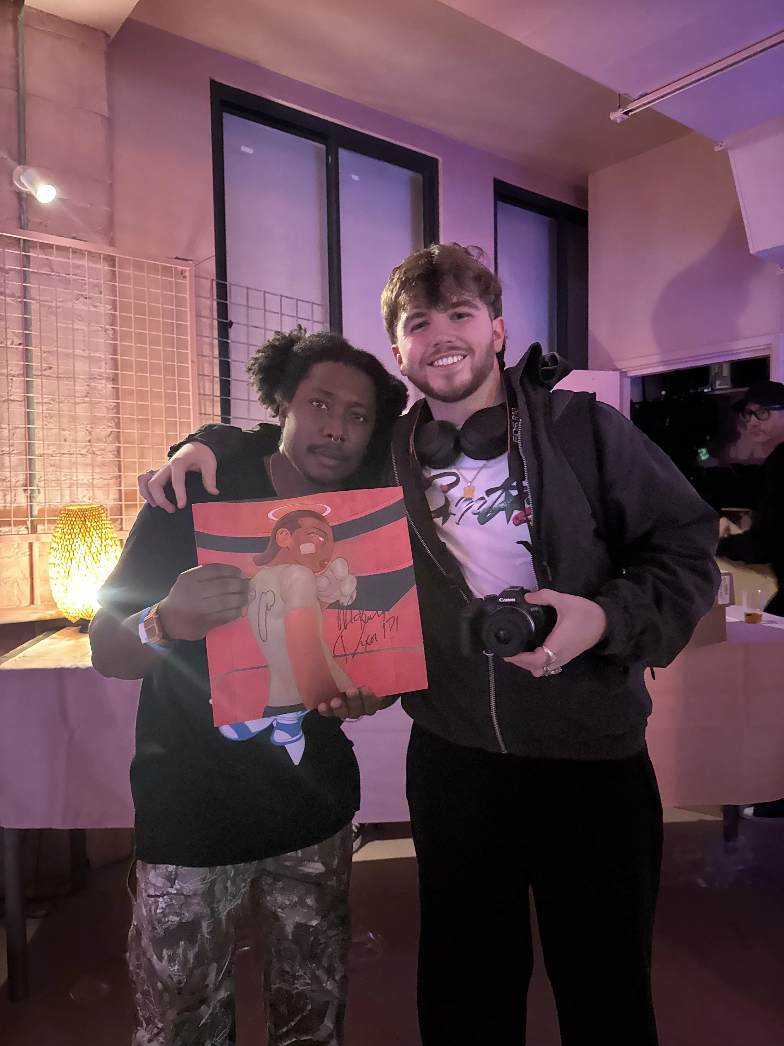 Two people posing together indoors, one holding a colorful caricature drawing of an angel with long hair, a halo, and wings, signed by the artist, with lighting and background suggesting an event or gathering.