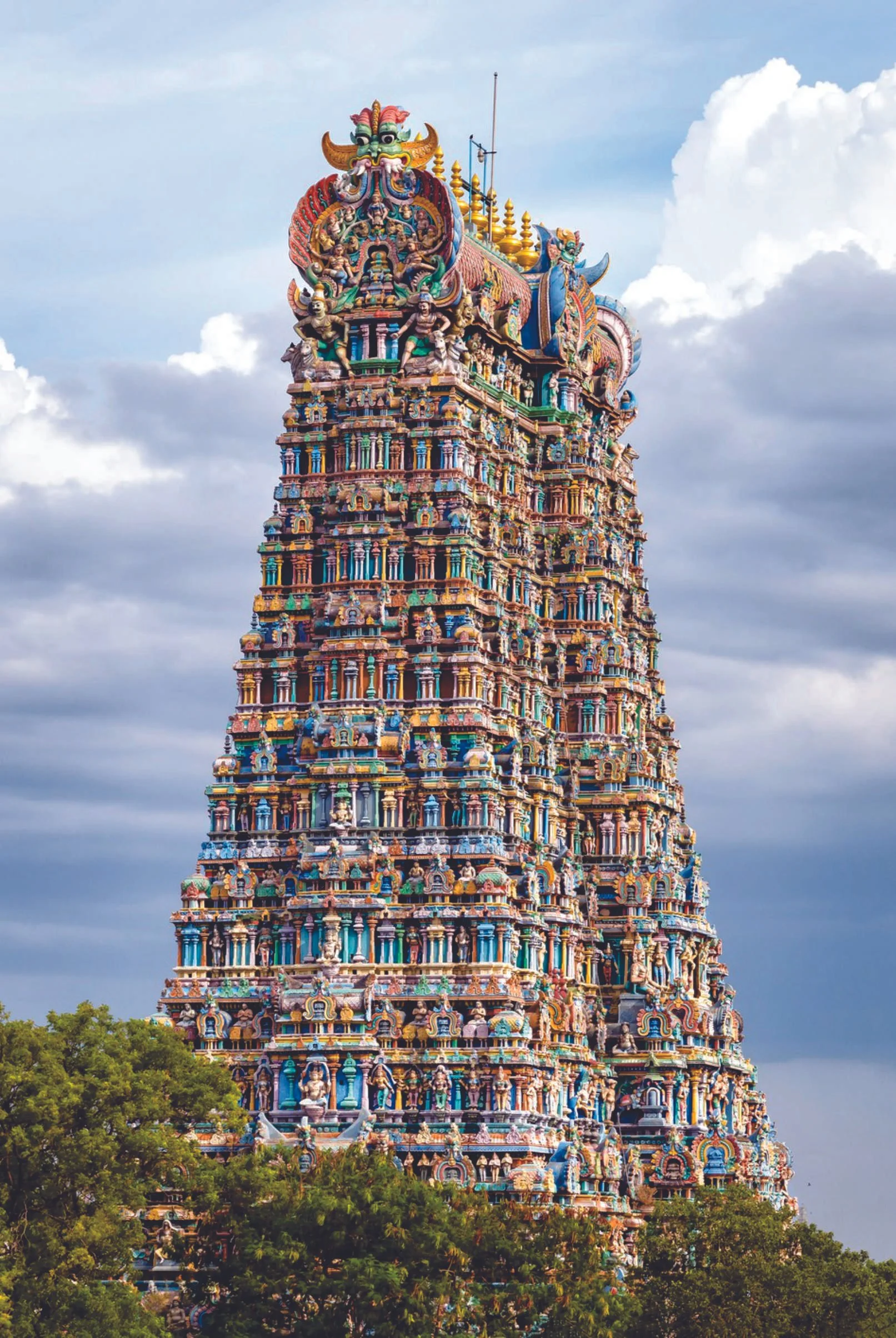 Colorful Hindu temple tower with intricate carvings and sculptures, set against a cloudy sky, with green trees in the foreground.