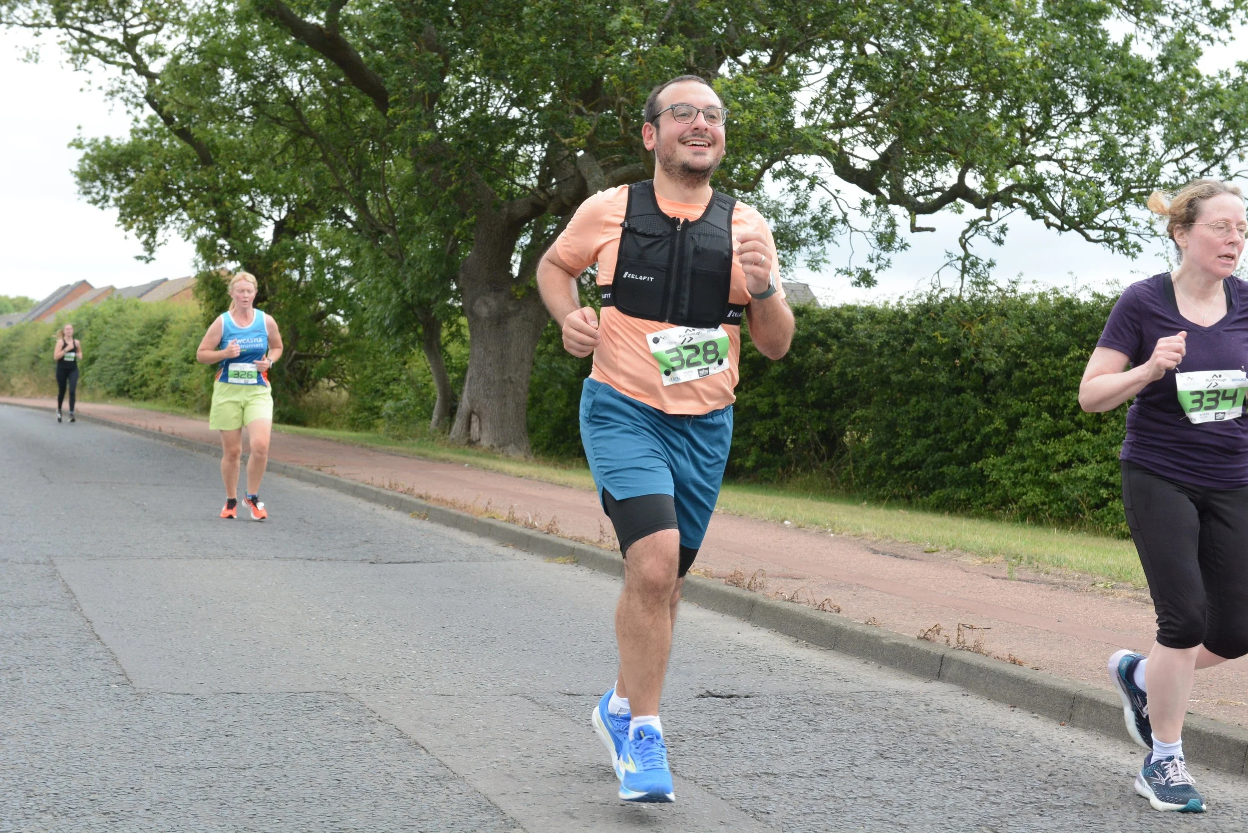 People participating in a road race or marathon, running on a paved street with greenery and trees in the background, dressed in activewear with race bibs.