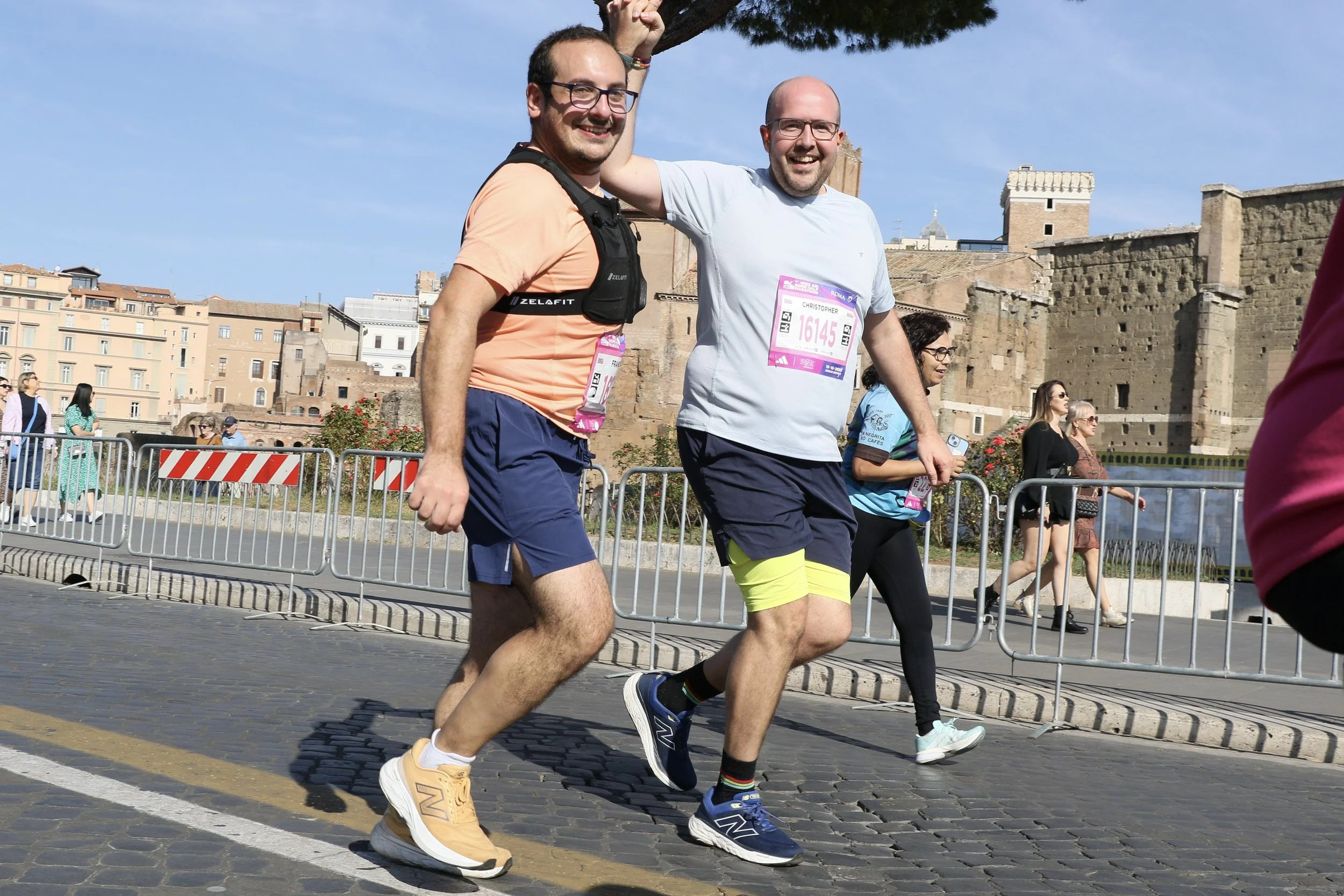 Two men running smiling during the Rome Half Marathon 2025, with race bibs, urban historic buildings in the background, and onlookers behind a metal barrier.