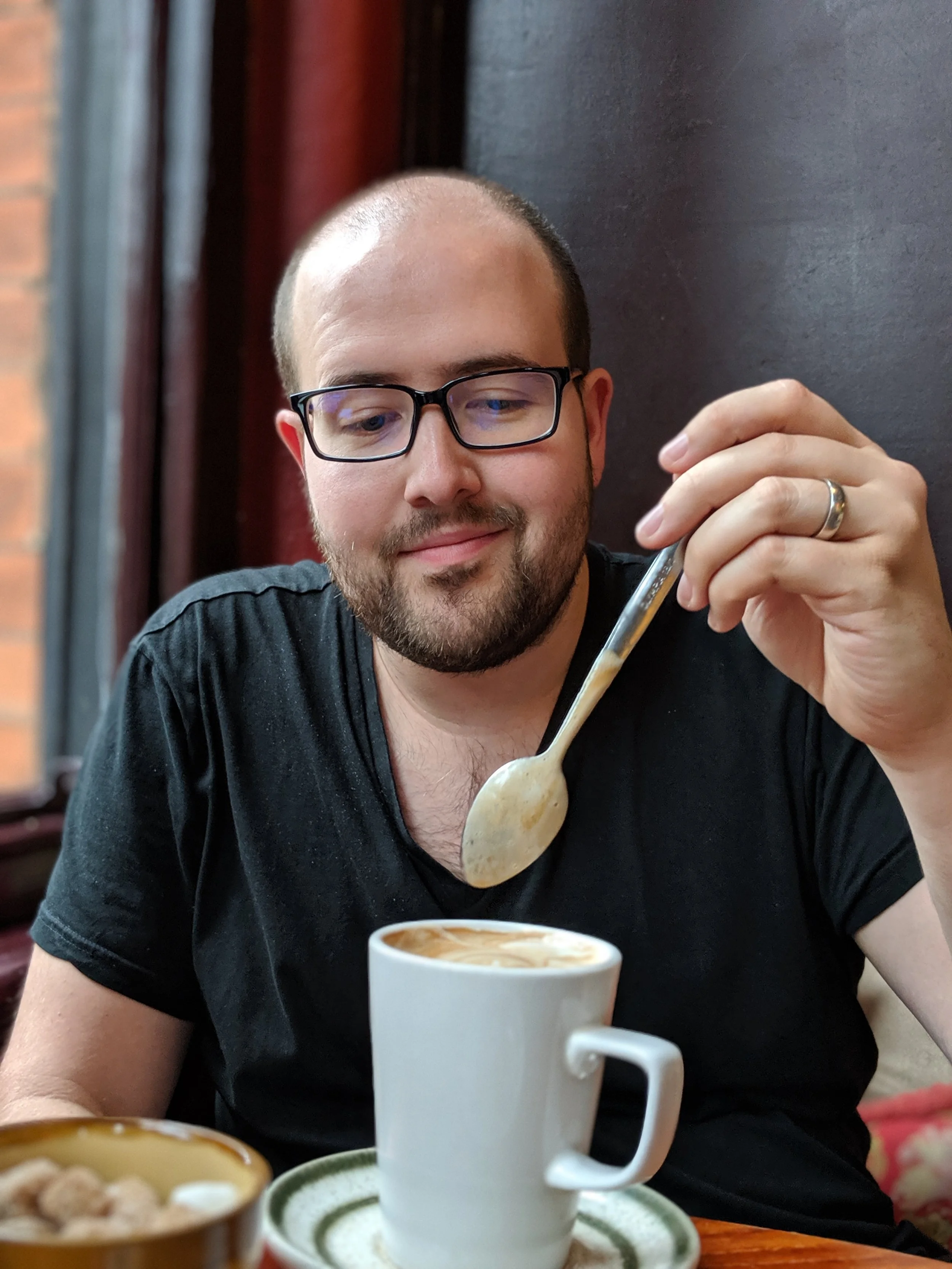 A man with glasses and a beard is stirring a latte in a white mug while sitting at a table in a cozy cafe.