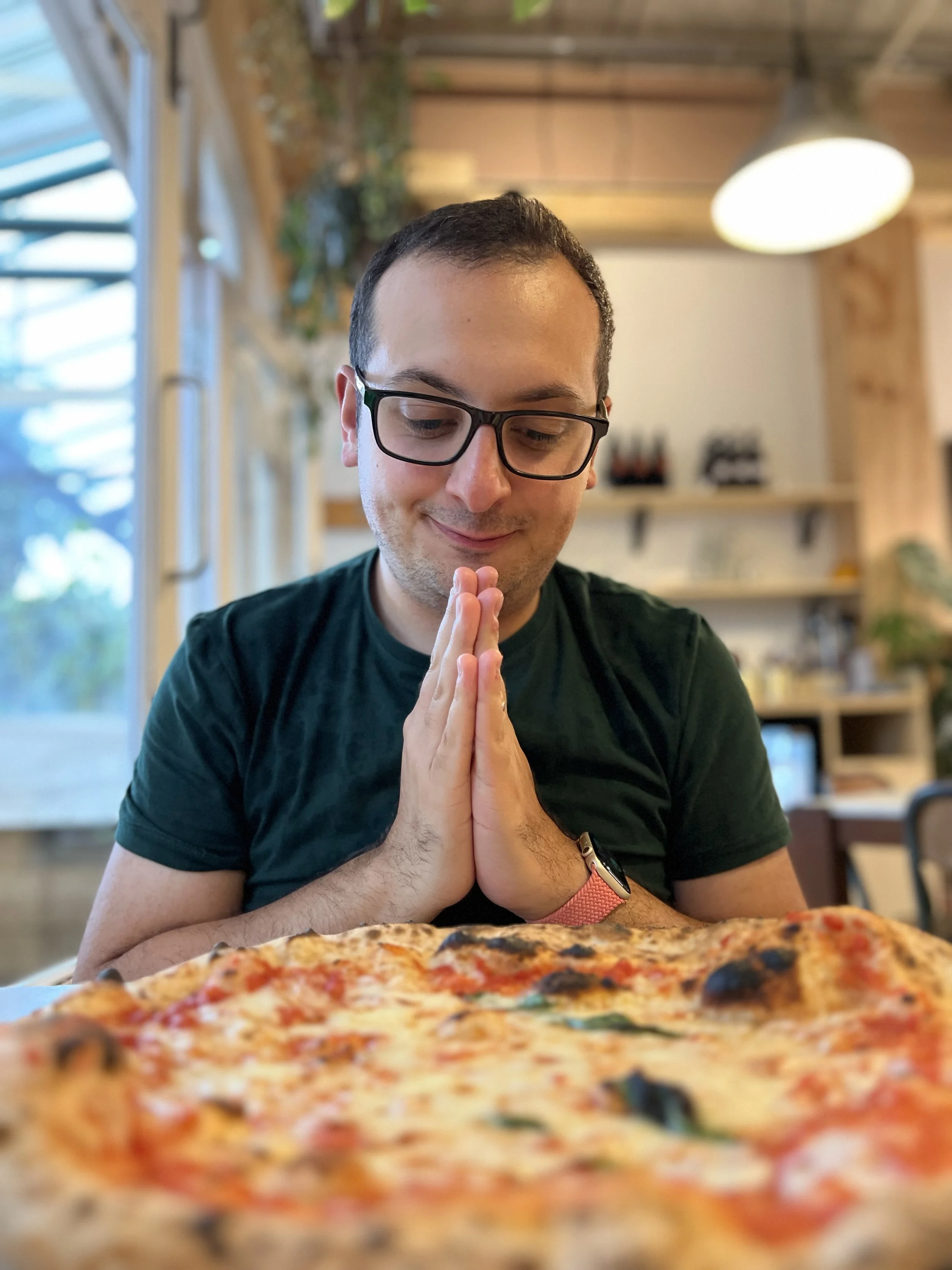 A man with glasses and a black t-shirt praying or giving thanks in front of a pizza in a cozy restaurant.
