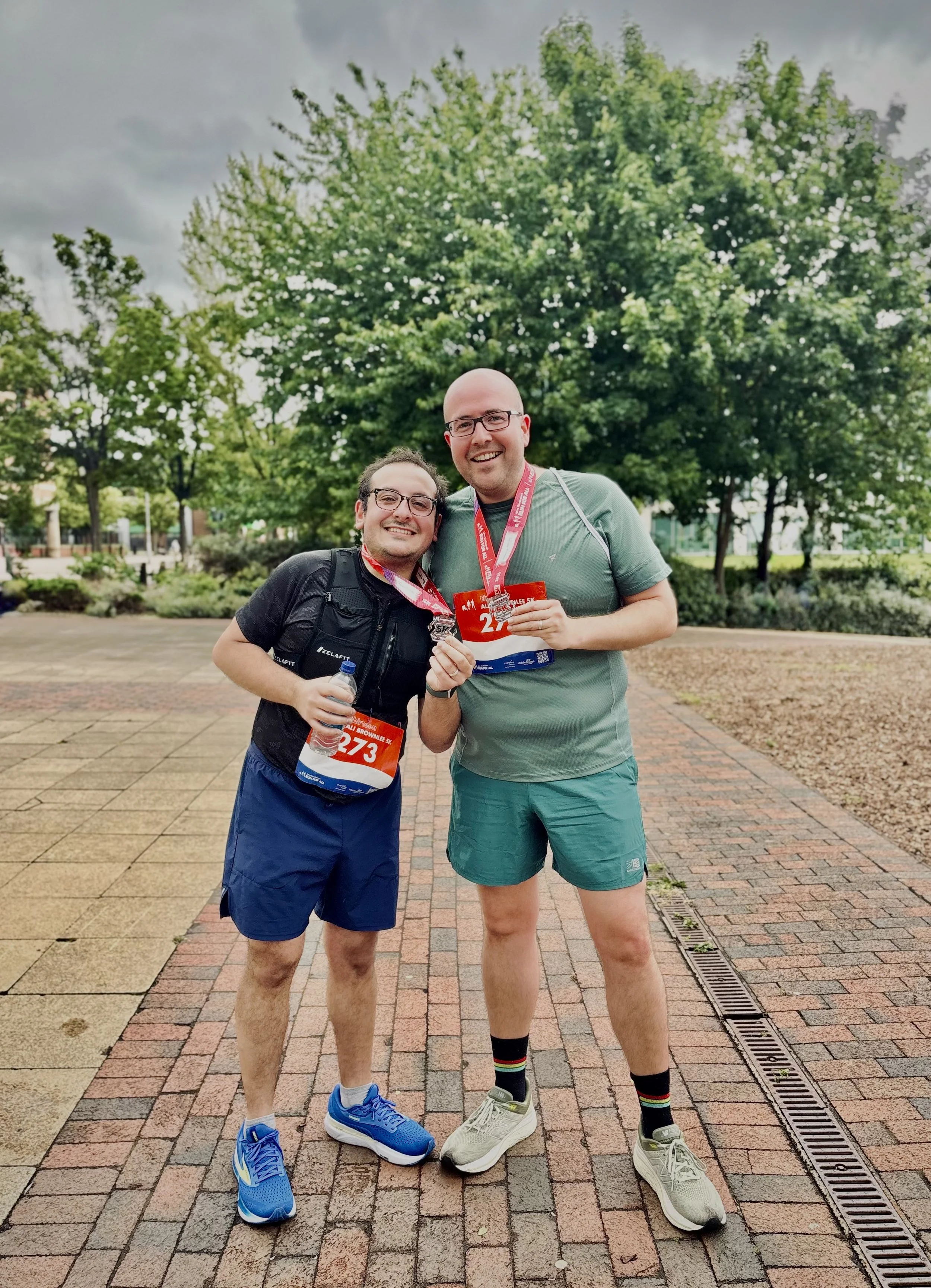 Two men standing outdoors after completing a race, wearing medals and race bibs, smiling at the camera. One is holding a water bottle, and the other is holding a race medal. The background features green trees and a cloudy sky.