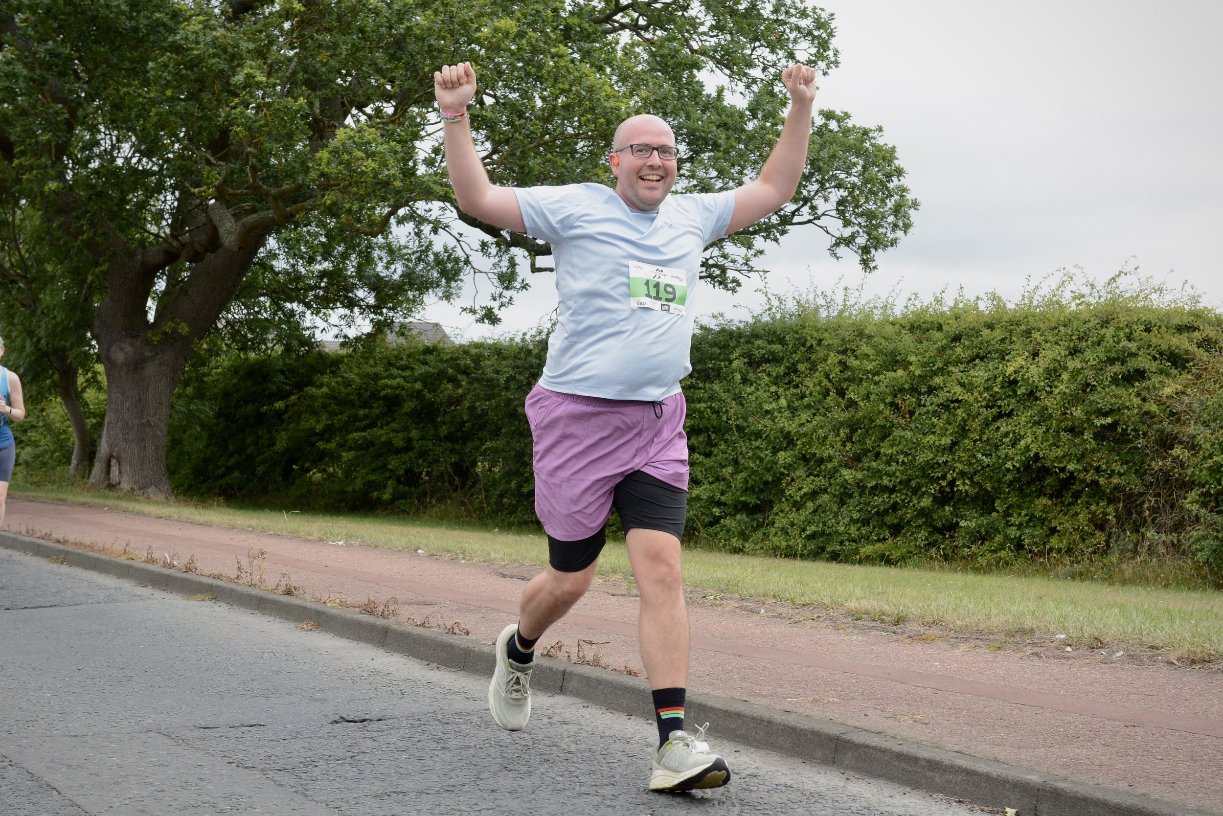A man running in a marathon, wearing a light blue shirt, purple shorts, black leggings, and white running shoes, with a race number '119' on his chest, raising his arms in celebration.