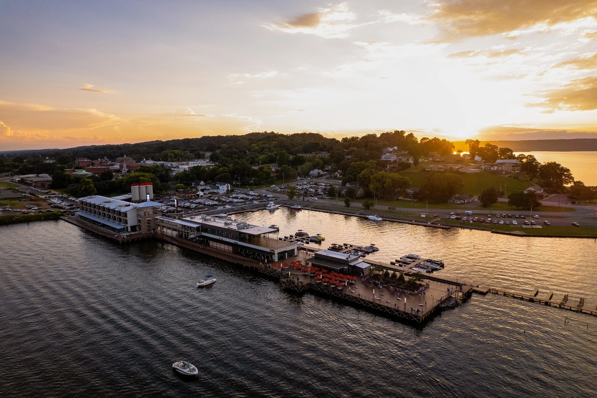 Aerial view of a waterfront restaurant with outdoor seating and boats docked in the river during sunset, with a hill and residential area in the background.