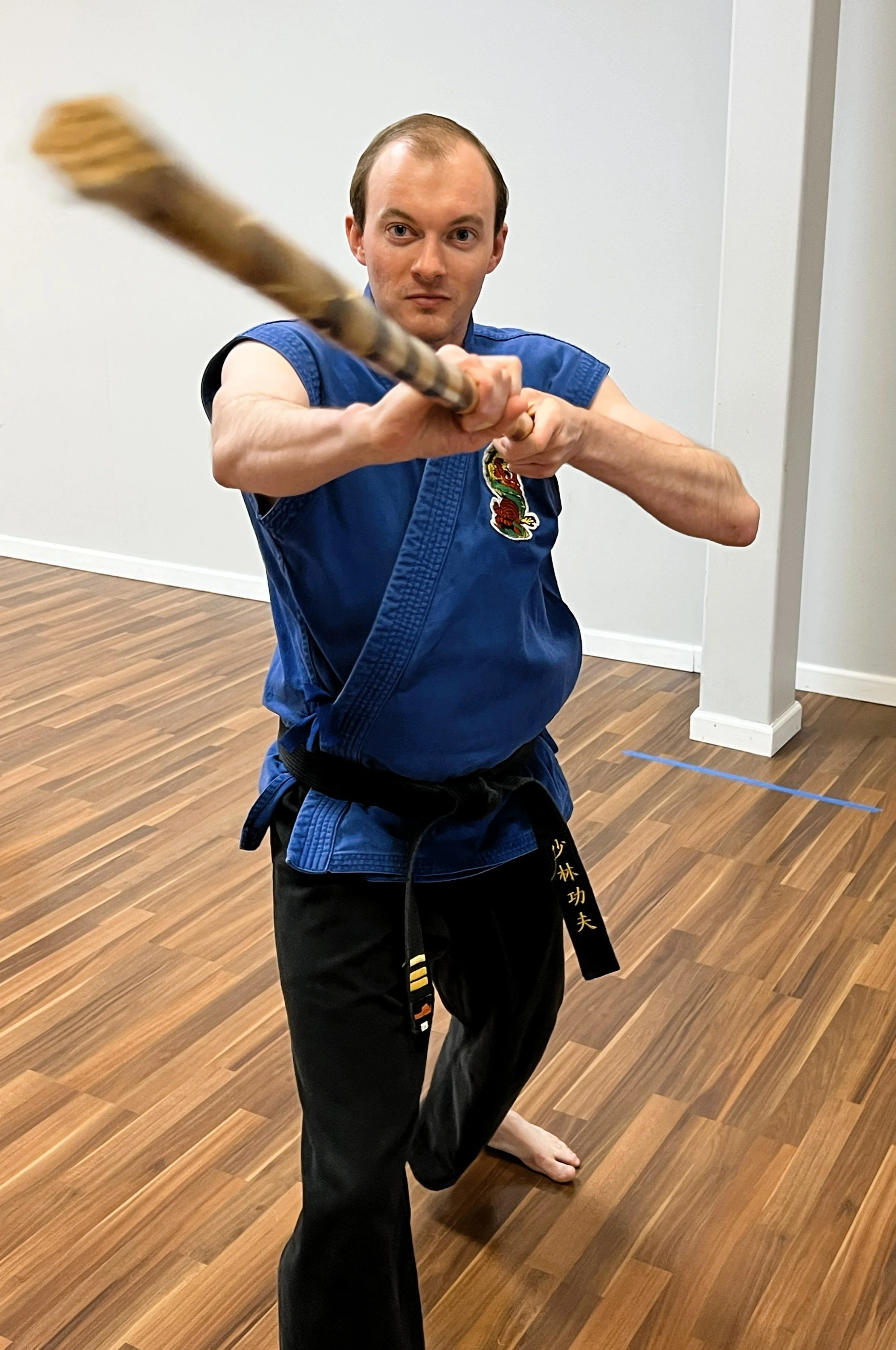 Man in martial arts uniform practicing with a staff in a studio with wooden floors.