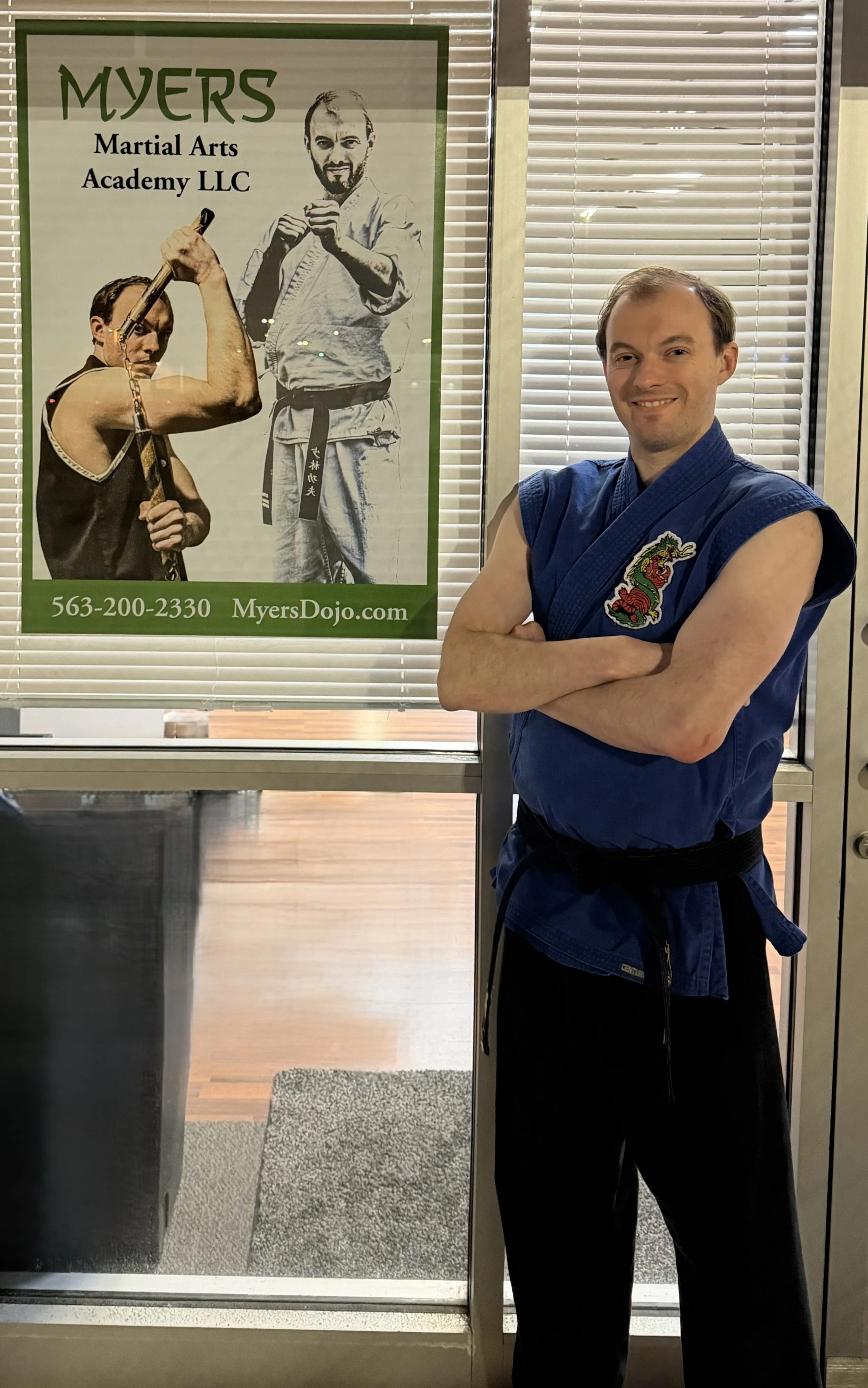 A man in a blue martial arts uniform with a dragon patch on his chest standing with arms crossed inside a martial arts studio. Behind him is a window with blinds and a poster for Myers Martial Arts Academy LLC with images of martial artists and contact information.
