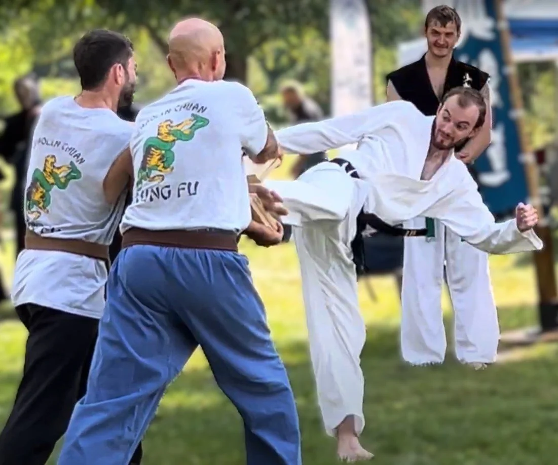Group of martial artists practicing or demonstrating martial arts outdoors, with one person in a white gi breaking two boards with his feet.
