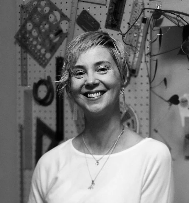 A smiling young woman with short, blonde hair, wearing a white top and layered necklaces, standing in a workshop with tools and equipment on a pegboard in the background.