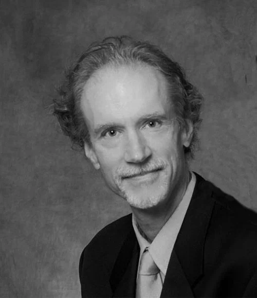 Black and white portrait of a middle-aged man with curly hair and a beard, wearing a suit and smiling.