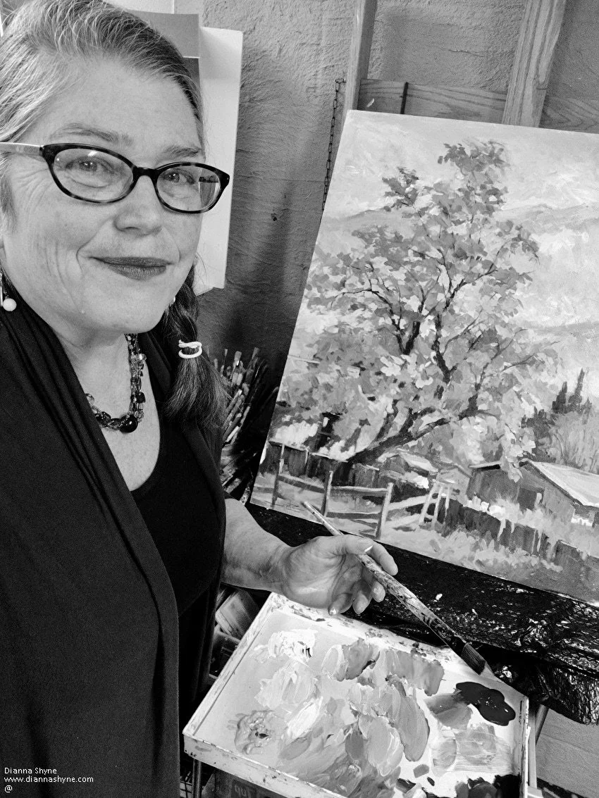A woman with glasses and jewelry, smiling, holding a paintbrush, standing next to an easel with a landscape painting of trees and fences, in an art studio.