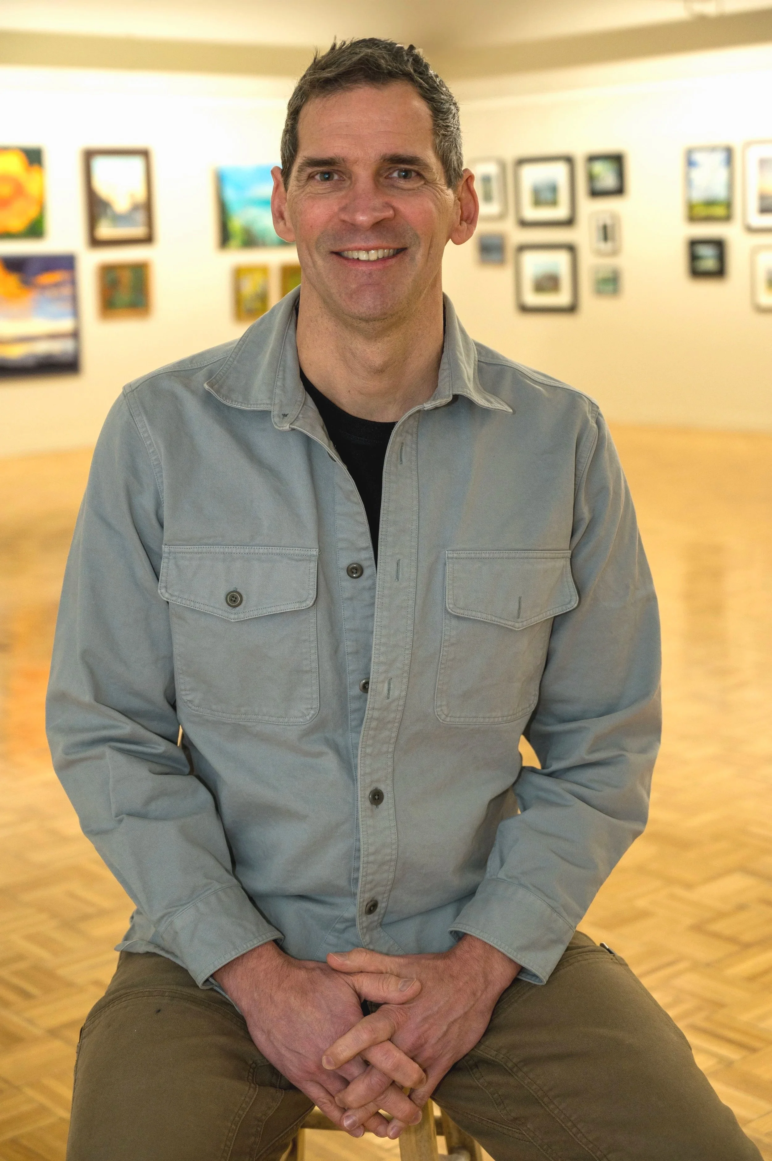 A man smiling while sitting on a stool in an art gallery with paintings on the wall behind him.