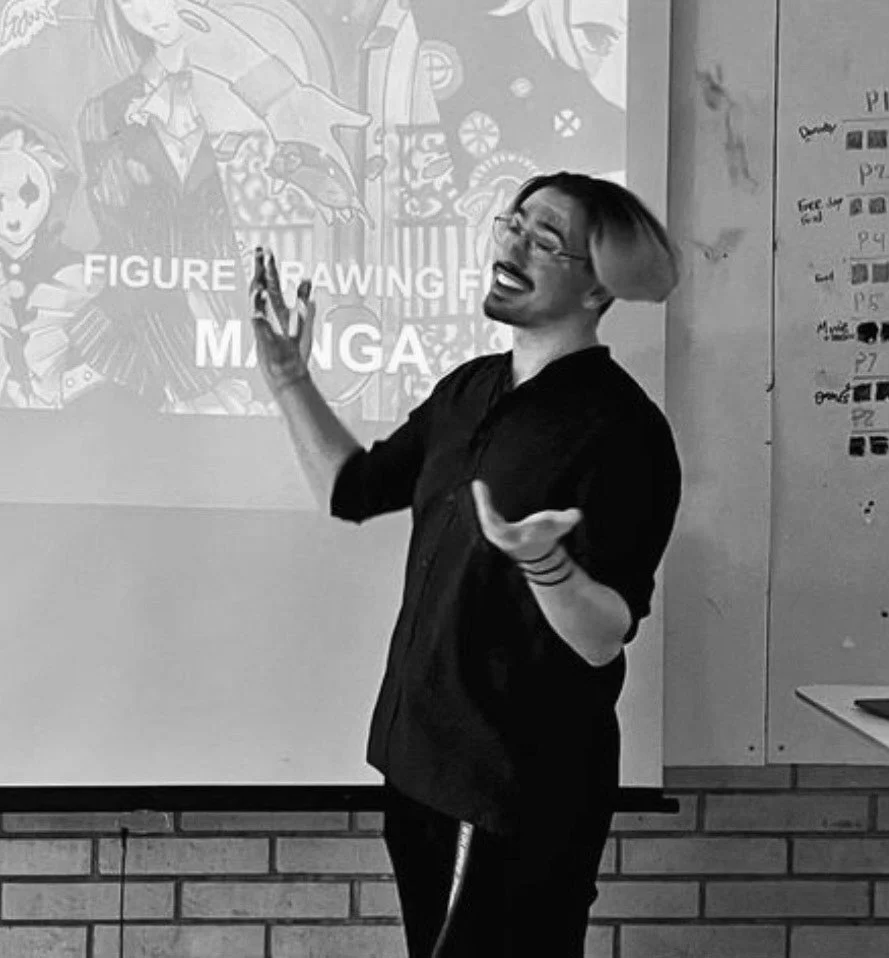 A woman with shoulder-length hair and glasses smiling and gesturing with hands while standing in front of a projected presentation slide about figure drawing manga.