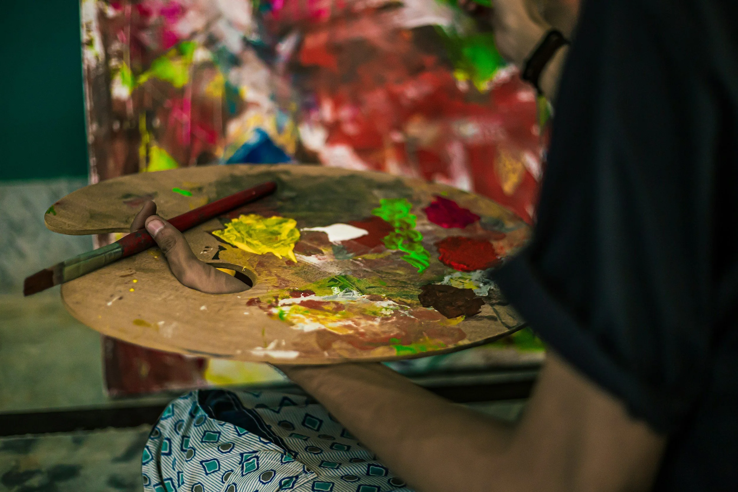 Person holding an artist's palette with various bright paint colors, in front of a colorful abstract painting.