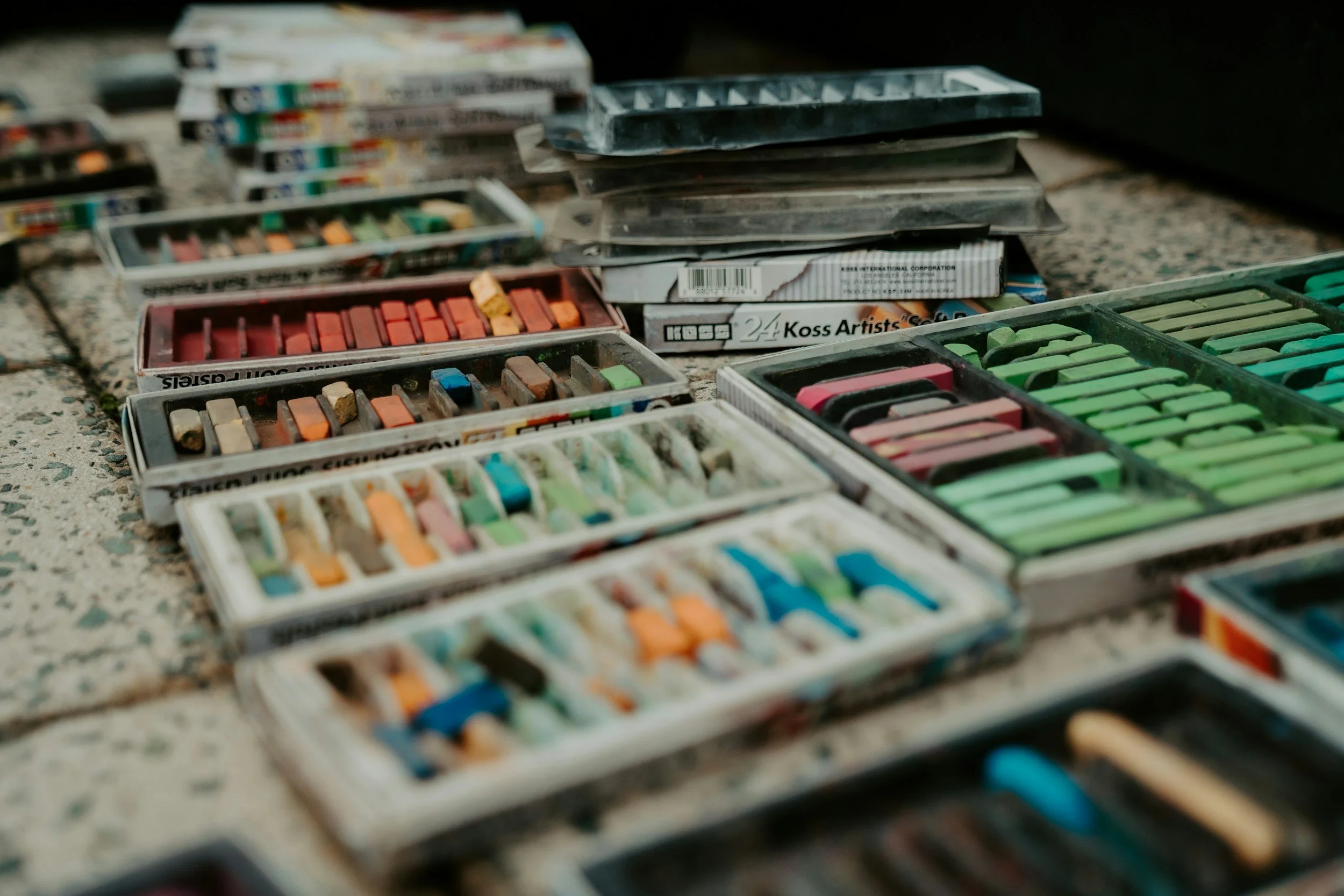 A collection of chalk pastels in various colors arranged in open boxes outdoors on a concrete surface.