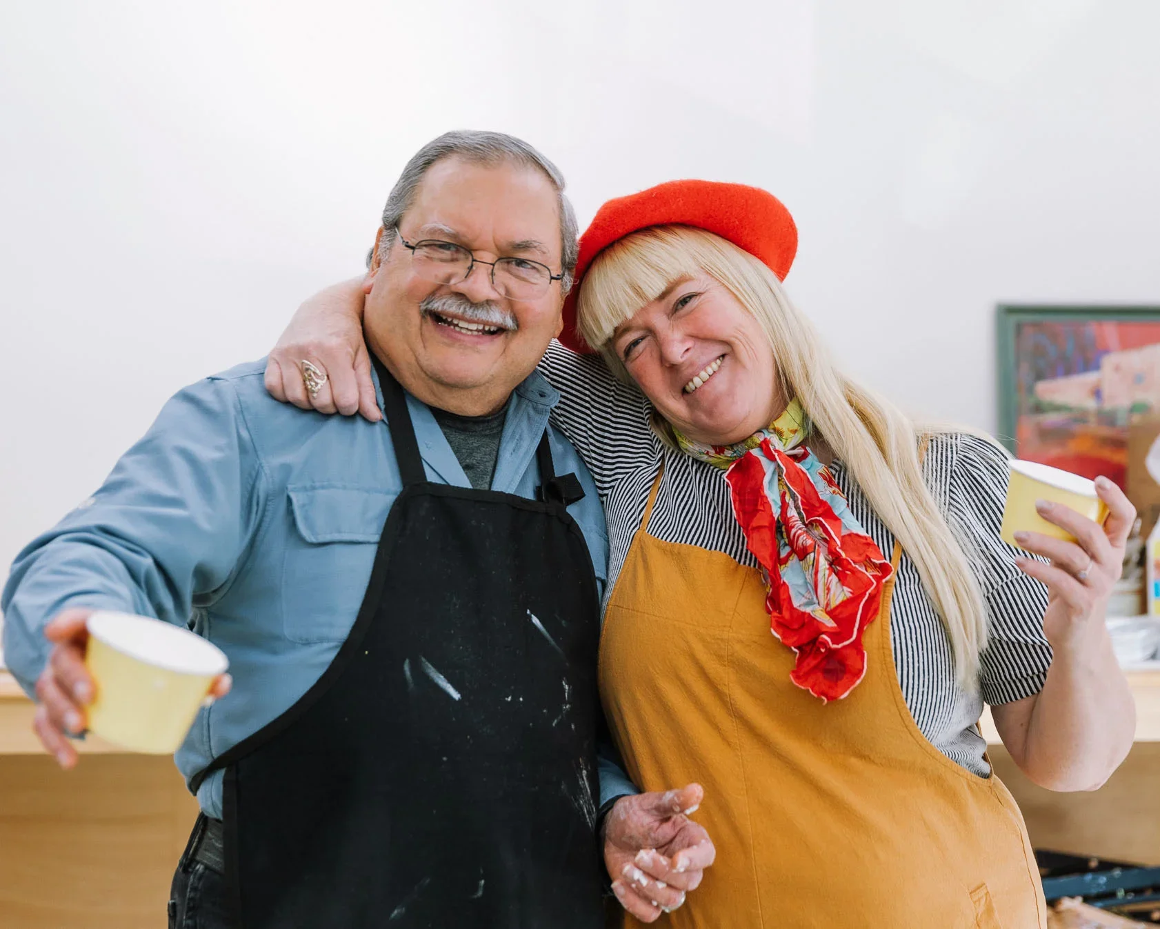 A smiling couple embracing, the man wearing glasses and an apron, and the woman wearing a red beret and a colorful scarf, holding cups in a cozy setting.
