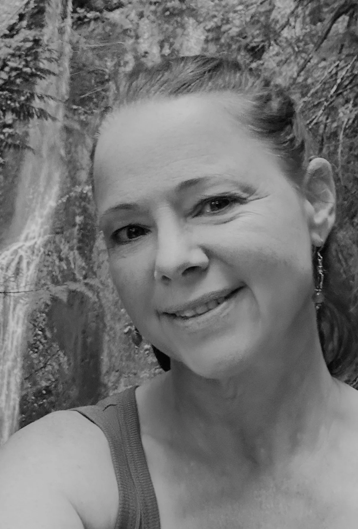 A woman smiling outdoors, with trees and a waterfall in the background.