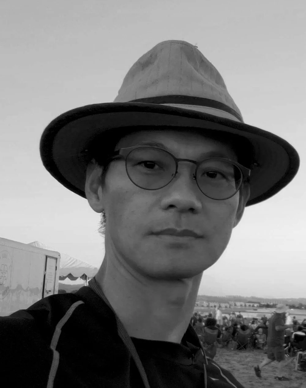 A man wearing glasses and a hat at an outdoor event with a crowd and tents in the background, in black and white.