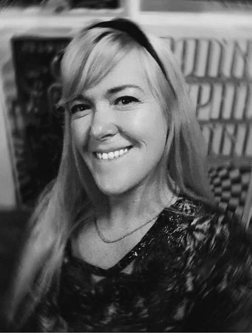 A smiling woman with long blonde hair and a headband, wearing a patterned top and a necklace, in a setting with signs and posters in the background.