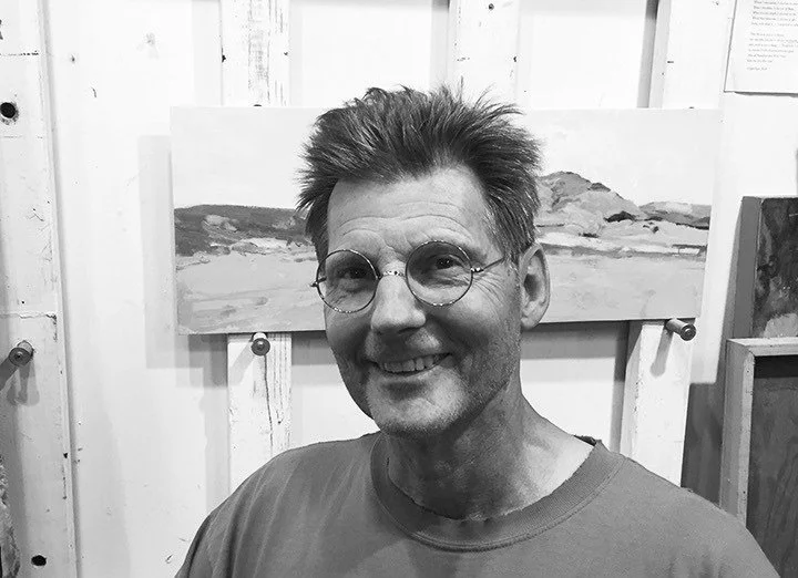 A smiling man with glasses and spiky hair in an art studio, with paintings of landscapes in the background.