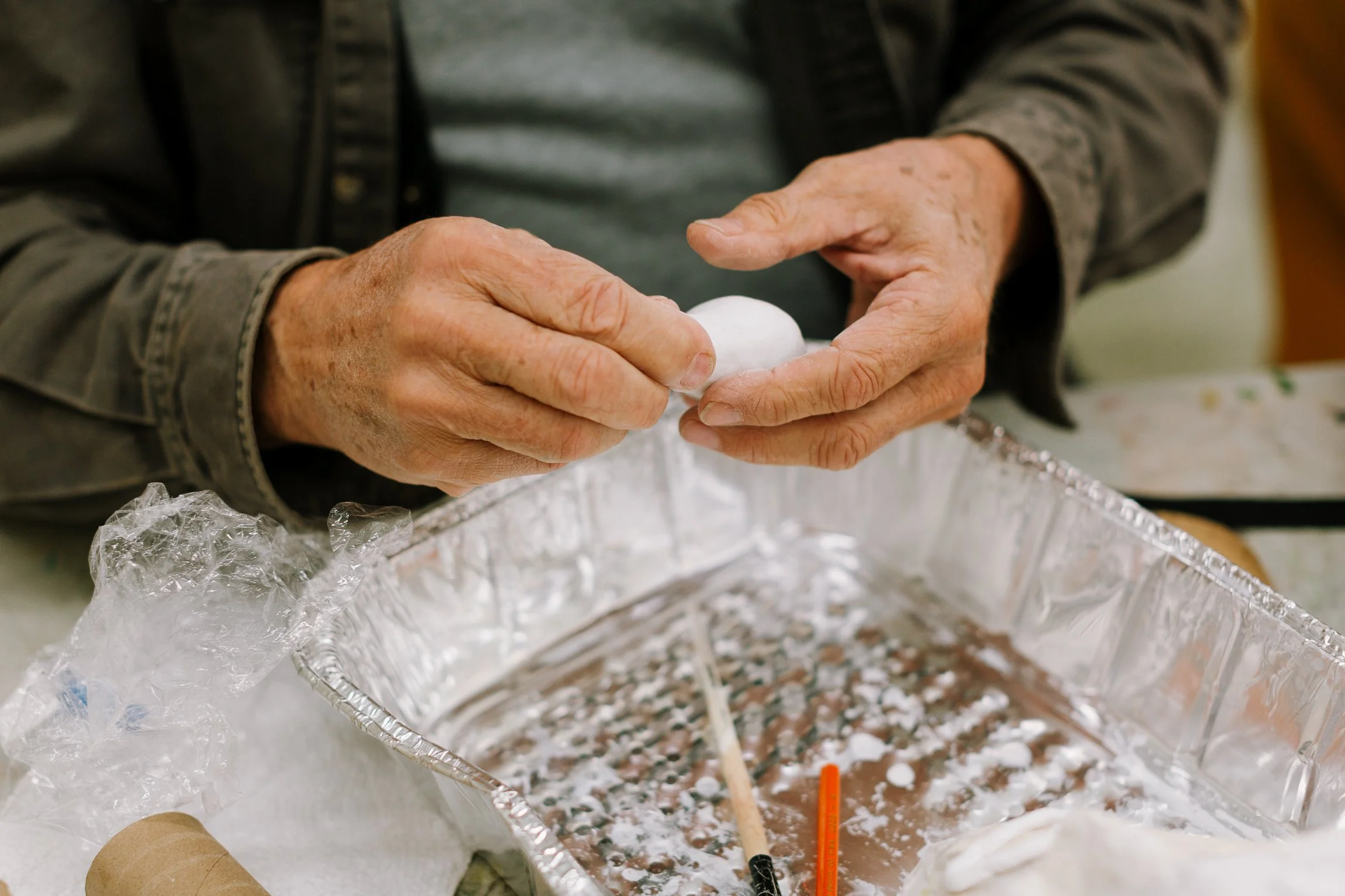Older person shaping a white object, possibly clay or soap, with their hands over a silver foil tray filled with similar material. Various tools and plastic wrap are on the tray.