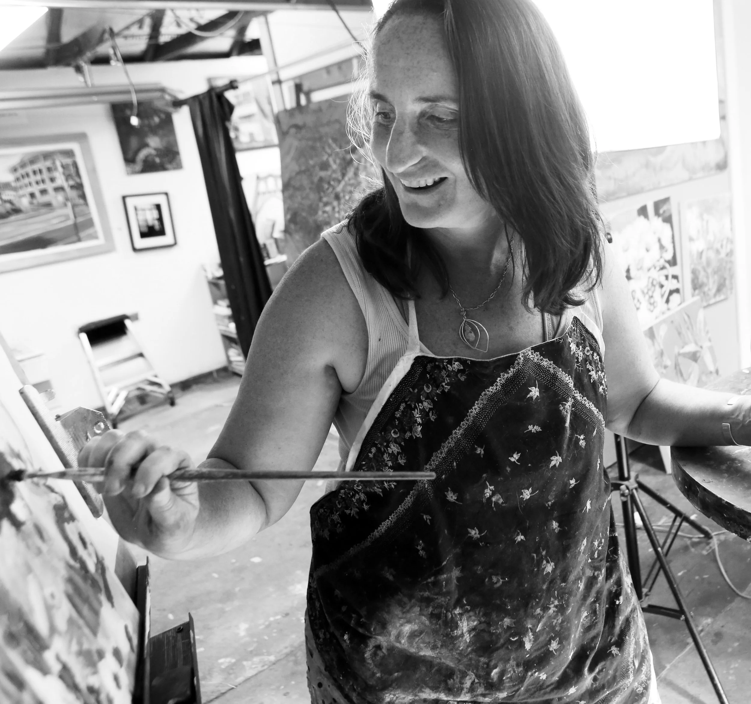 A woman painting on a canvas in an art studio, wearing an apron and smiling.