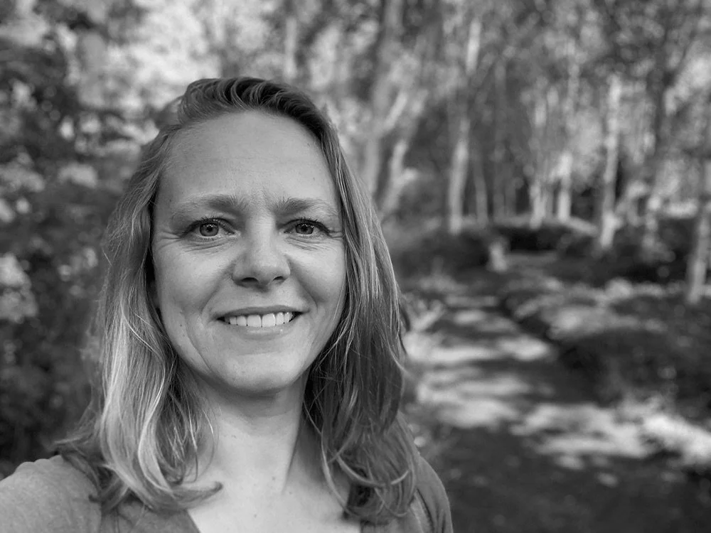 A woman with shoulder-length hair taking a selfie outdoors on a trail, with trees and foliage in the background.