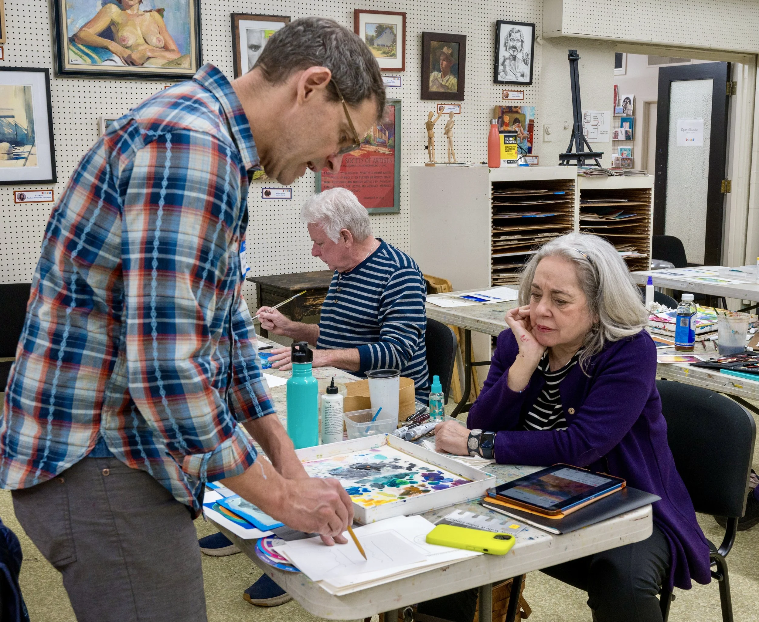 Two women and a man in an art class, with one woman seated at a table considering her artwork, another woman working at a separate table, and a man standing at a table using a paintbrush, surrounded by art supplies and artworks on the wall.