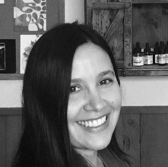 Smiling woman with long dark hair in a black and white photo, with wooden shelves and bottles in the background.