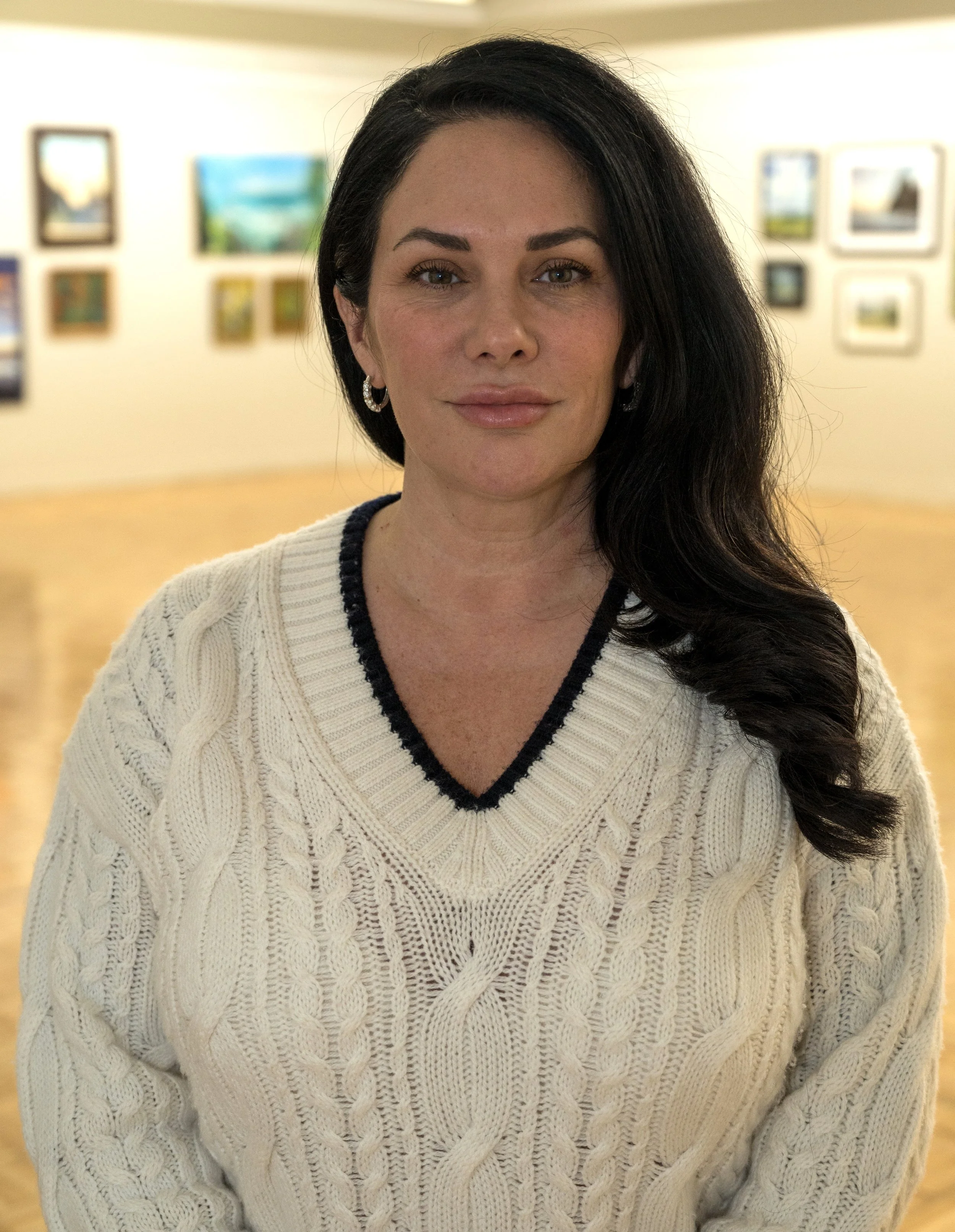 A woman with dark hair, wearing a beaded hoop earring and a cream-colored cable knit sweater with a black-edged V-neck, stands in an art gallery with framed paintings on the wall behind her.
