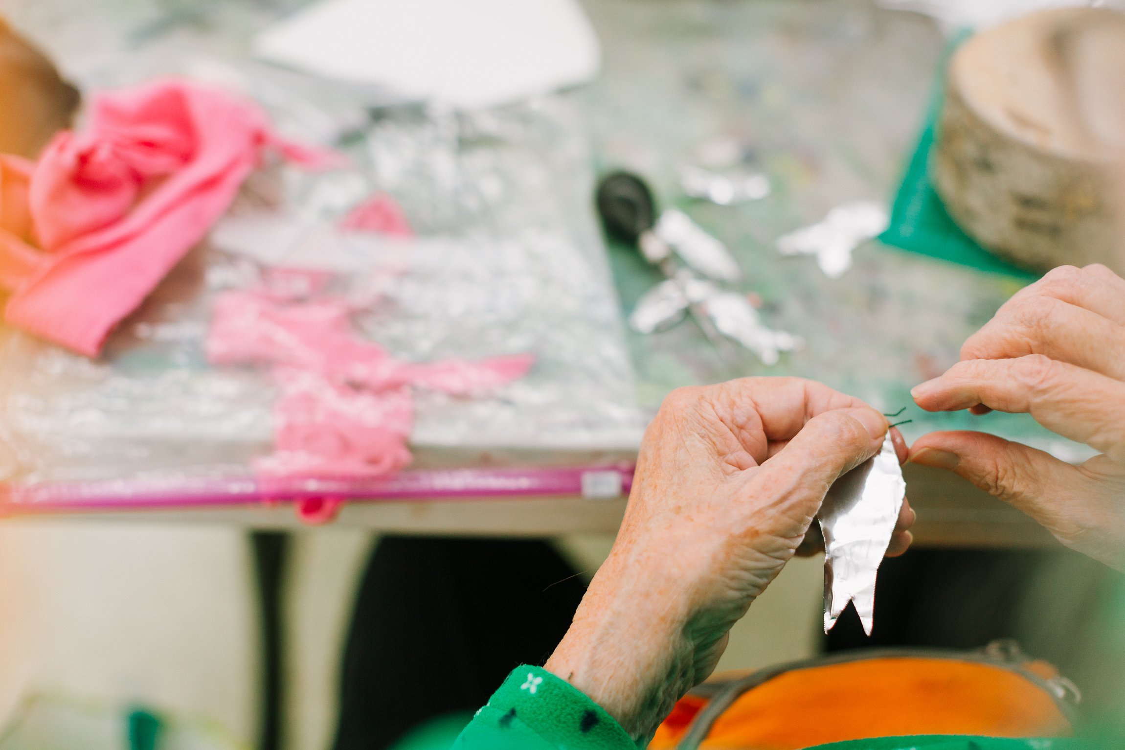 Close-up of elderly person's hands shaping silver foil into a small fish or leaf shape, with crafting supplies like pink fabric, scissors, shells, and tools in the blurred background on a work table.