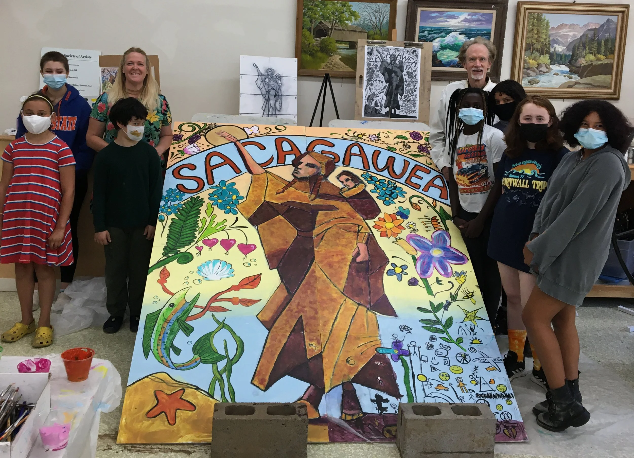 Group of children and adults standing around a colorful mural with the word 'SACAGAWEA' and a depiction of Sacagawea carrying her baby, surrounded by flowers, animals, and symbols, in an art studio with paintings on the walls.