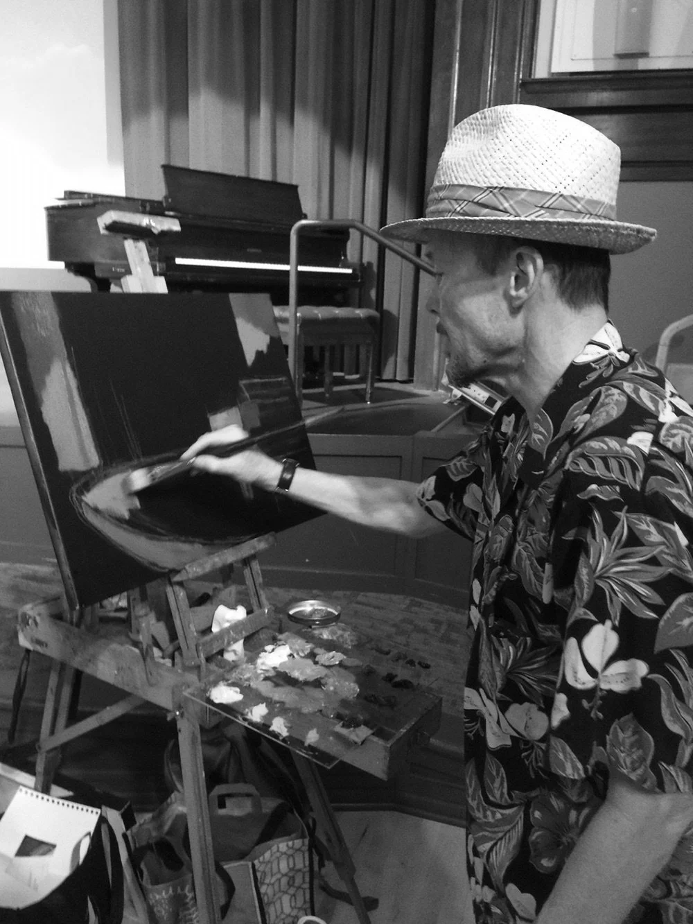 A man wearing a straw hat and a Hawaiian shirt is painting on a canvas in an art studio.