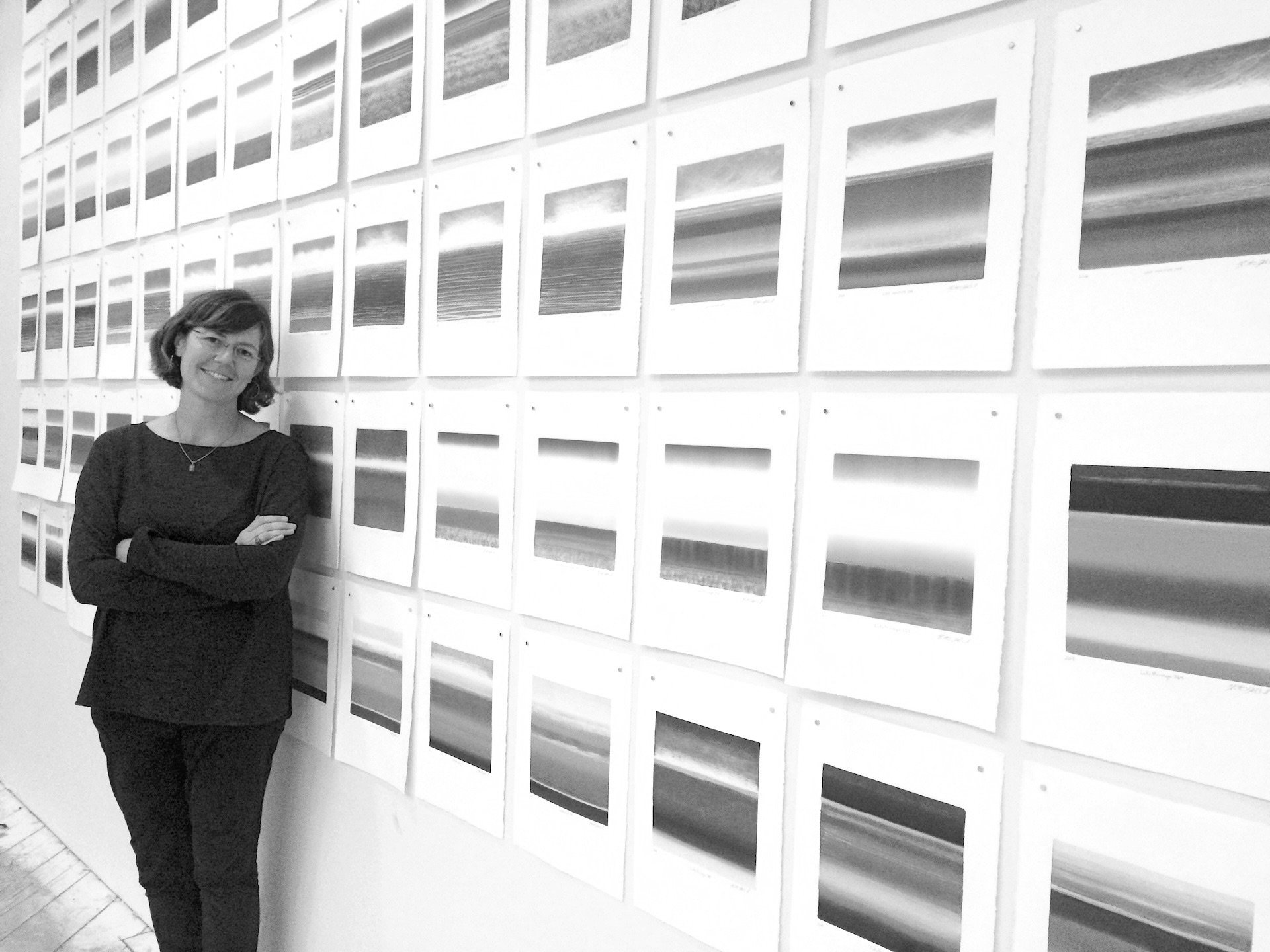 Woman standing with arms crossed in front of a wall of framed abstract art pieces.