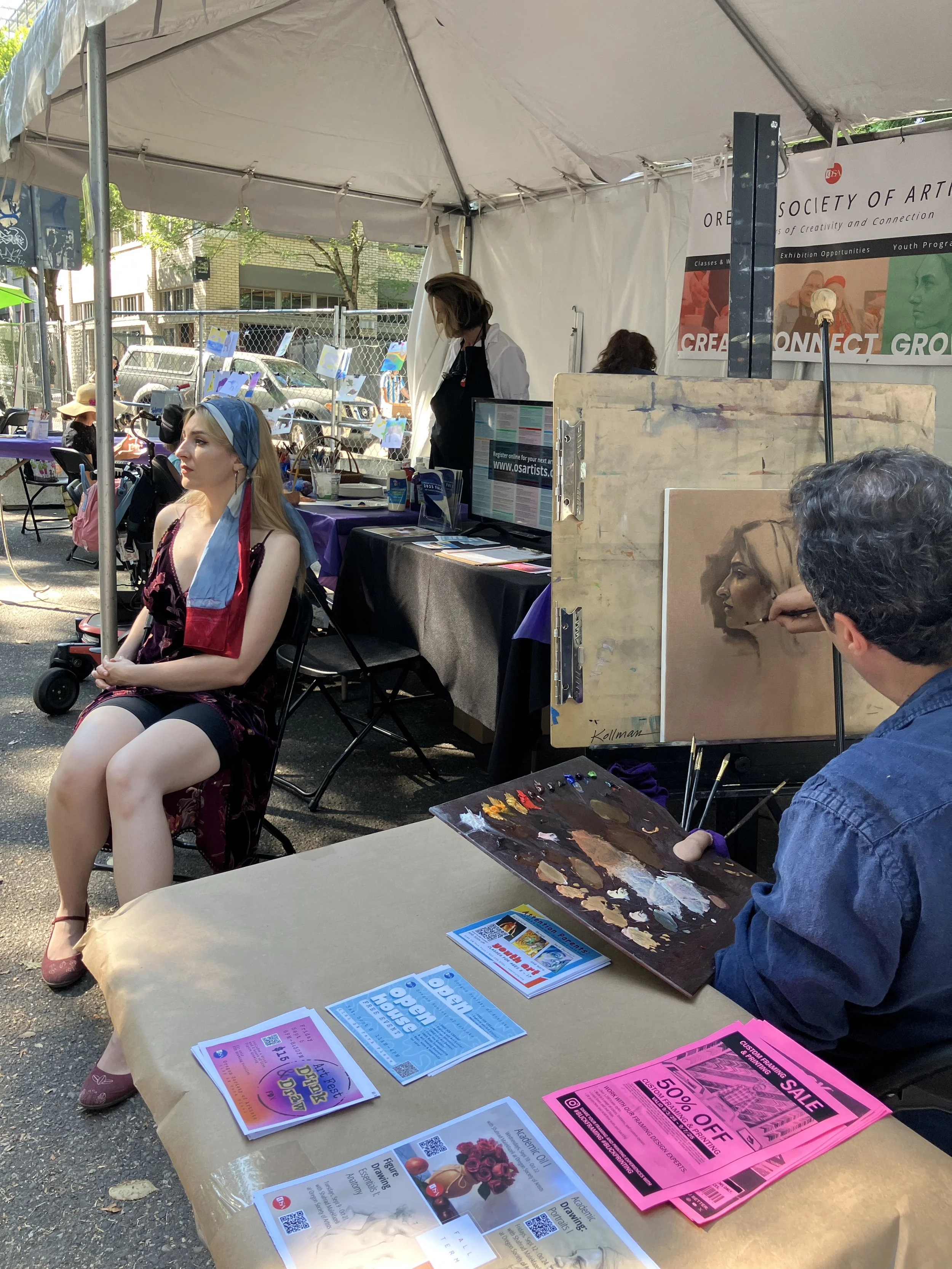 An outdoor art booth with a woman painting a portrait of a woman with blonde hair. The booth is under a white tent, and there are promotional flyers on the table in front. The seated woman has a blue and red scarf around her head and is dressed in a dark dress. There is a person behind the artist working on their art.