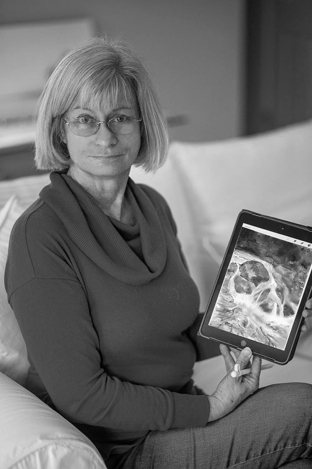 A woman with glasses and short hair sitting on a bed, holding a digital tablet displaying a landscape painting.