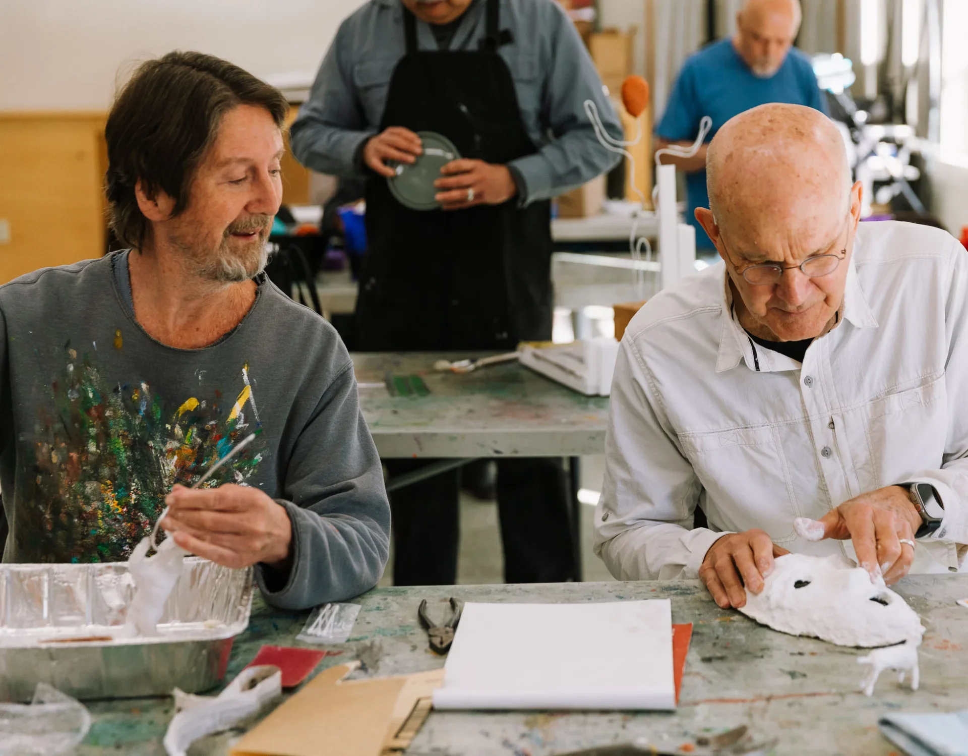 Two men working on a creative project at a table in a workshop. One is holding a paper or fabric mask, while the other is adding details to it. In the background, a person prepares food and another looks on.