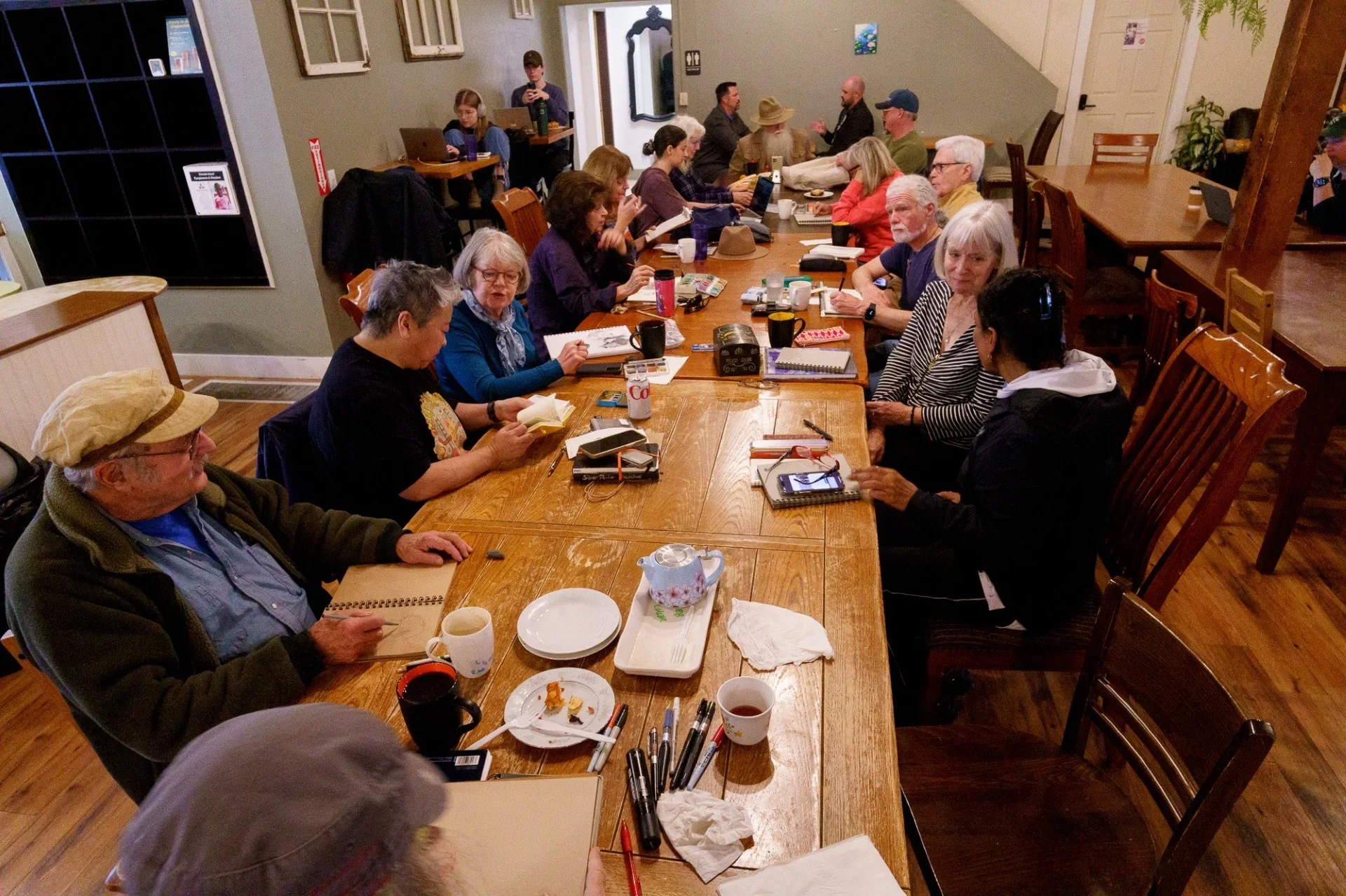 Group of people sitting around a large wooden table in a cozy indoor room, engaged in reading, writing, and conversation.