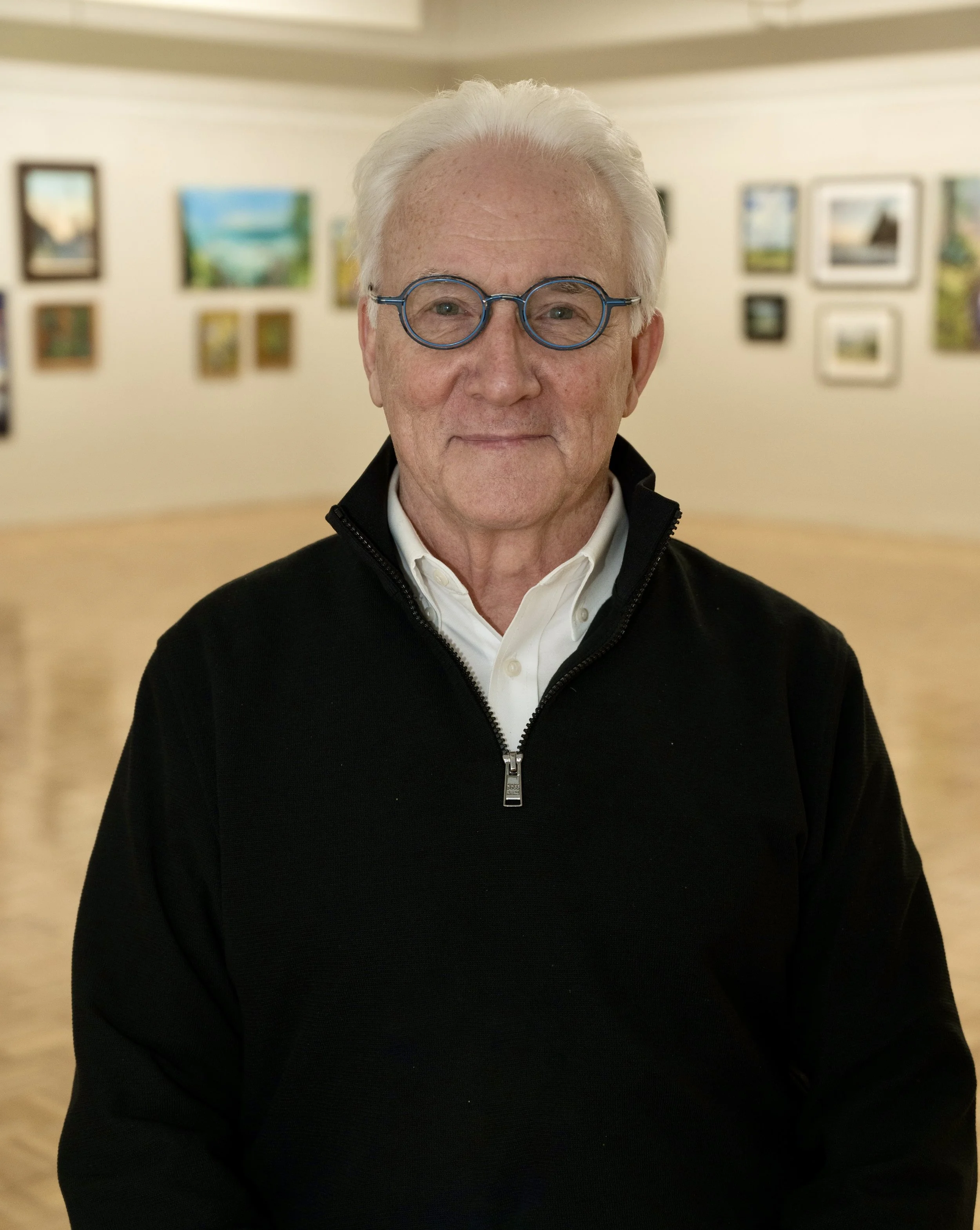 An elderly man with white hair and glasses, wearing a white shirt and black zip-up jacket, standing in an art gallery with paintings on the wall behind him.