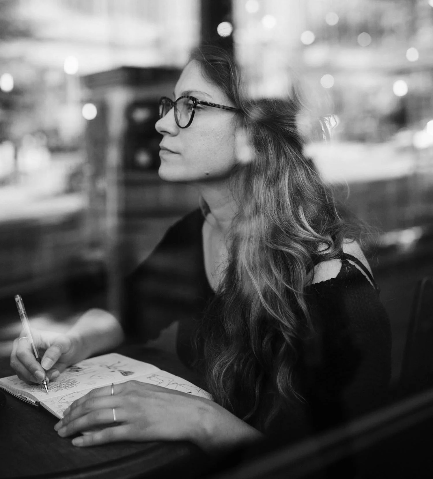 A woman with glasses and long wavy hair sitting at a table, writing or drawing in a notebook, with a blurred cityscape background visible through a window.