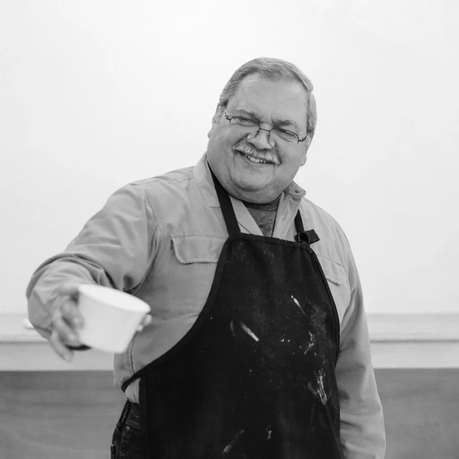 A smiling man wearing glasses, a light-colored shirt, and a dark apron, holding a small bowl, standing in a plain room.