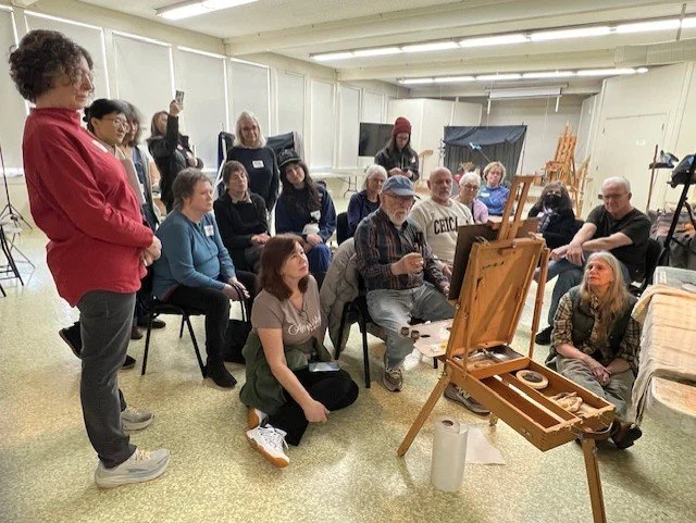 A group of people gathered in an art studio or classroom, some seated and some standing, watching an artist demonstrate painting on an easel, with painting supplies and seated viewers nearby.