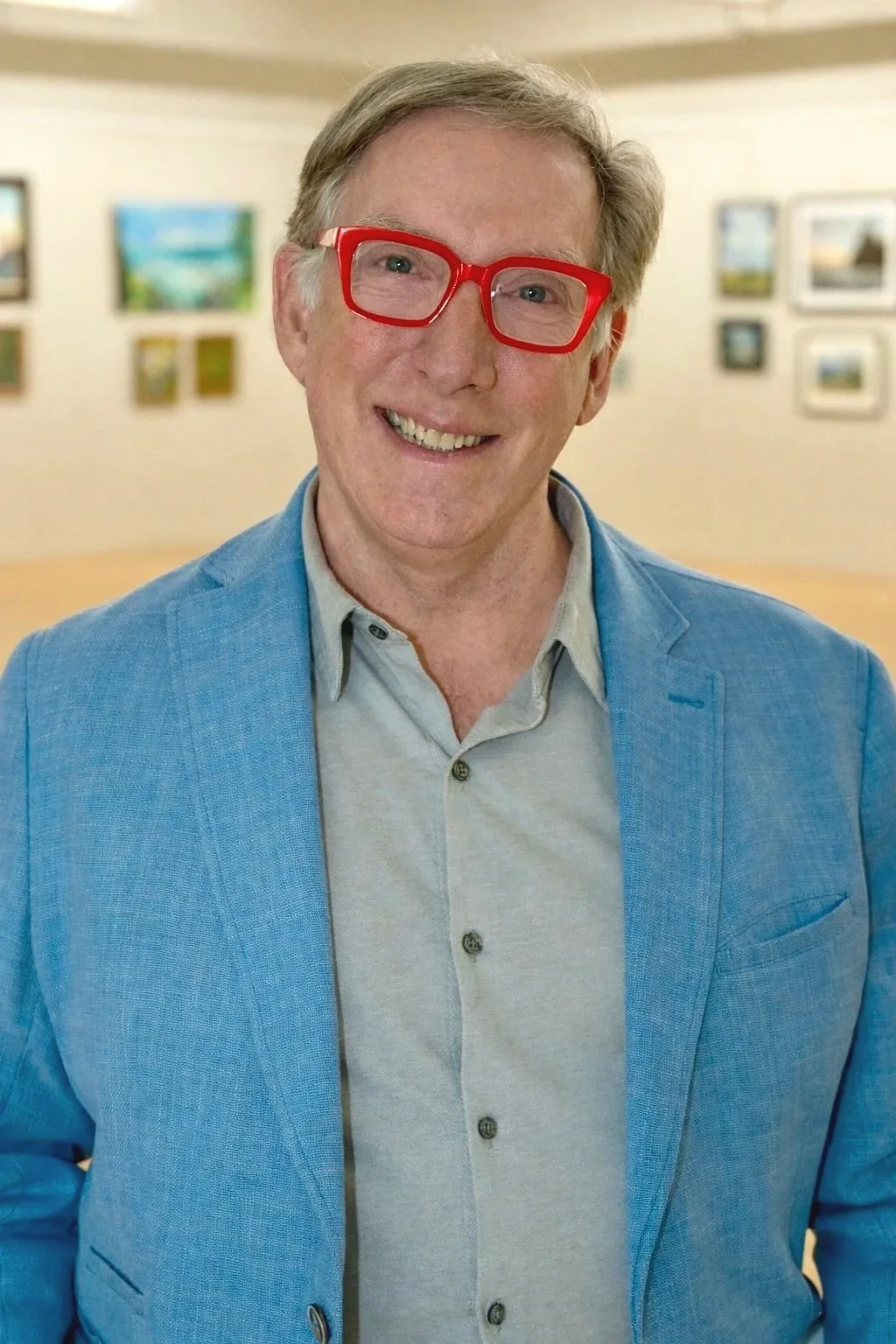 A smiling man with red glasses, wearing a blue blazer and a light gray button-up shirt, standing in an art gallery with framed pictures on the wall.