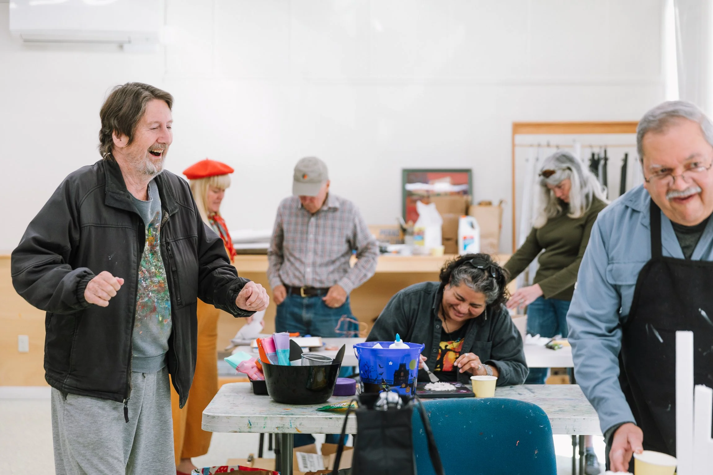A group of adults at a craft or painting workshop, smiling and interacting in a well-lit room.